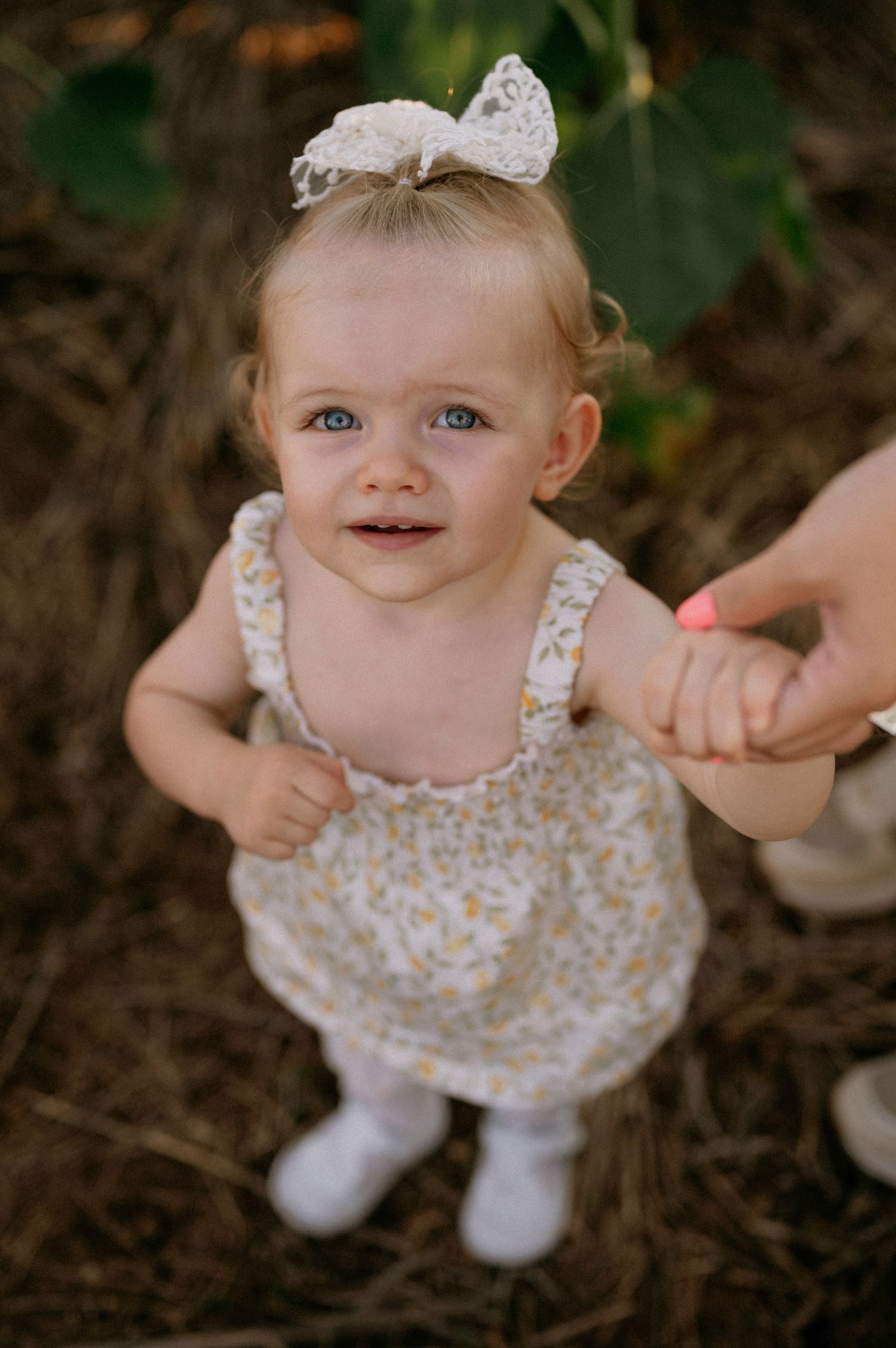 Adorable baby girl in floral romper and white bow headband holds an adult hand outdoors.