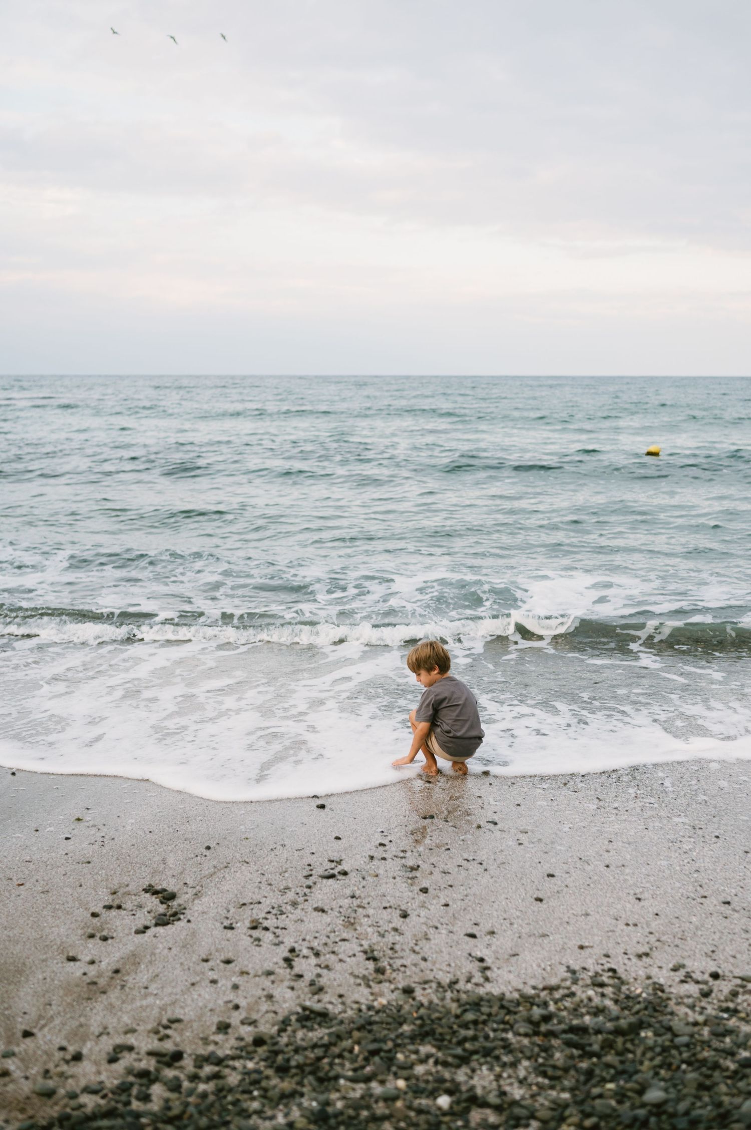Young child playing at the ocean shoreline, waves washing over sandy pebble beach on overcast day.