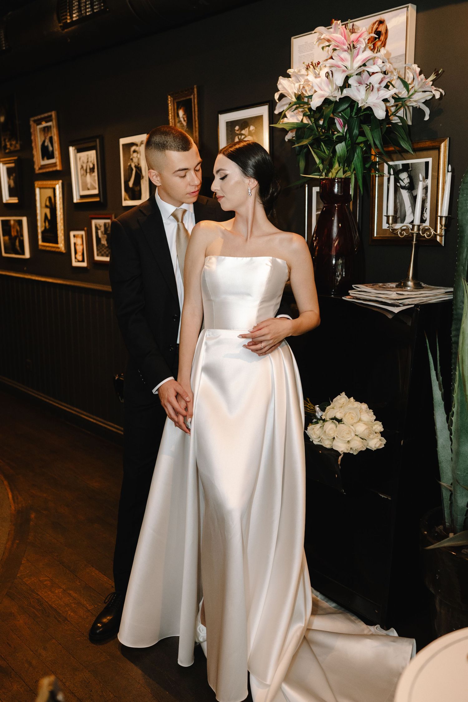 Romantic wedding couple in formal attire embracing on dark hardwood floor with framed artwork on walls.