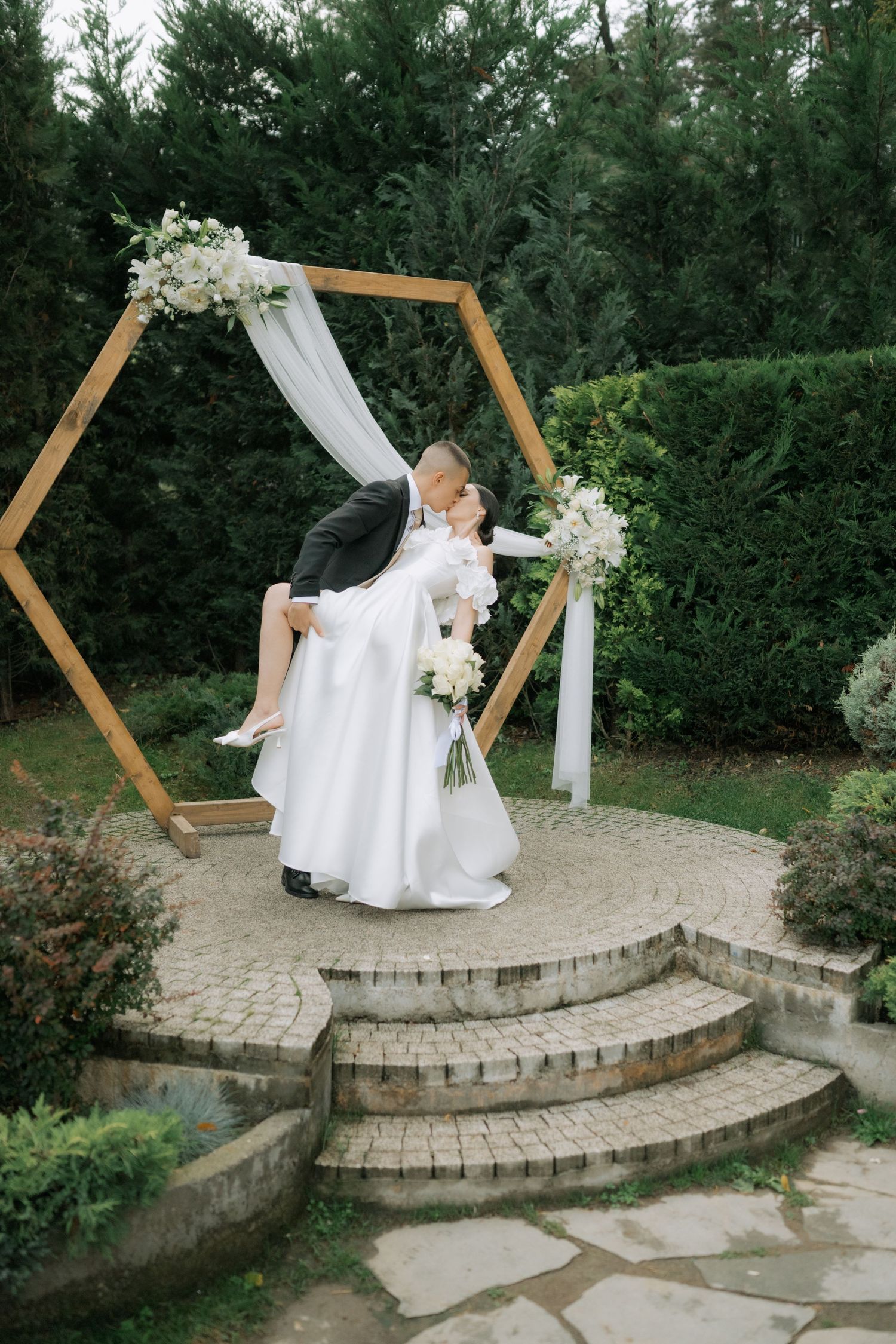 A series of outdoor wedding photos featuring a stone staircase decorated with a wooden hexagon arch and white drapery.