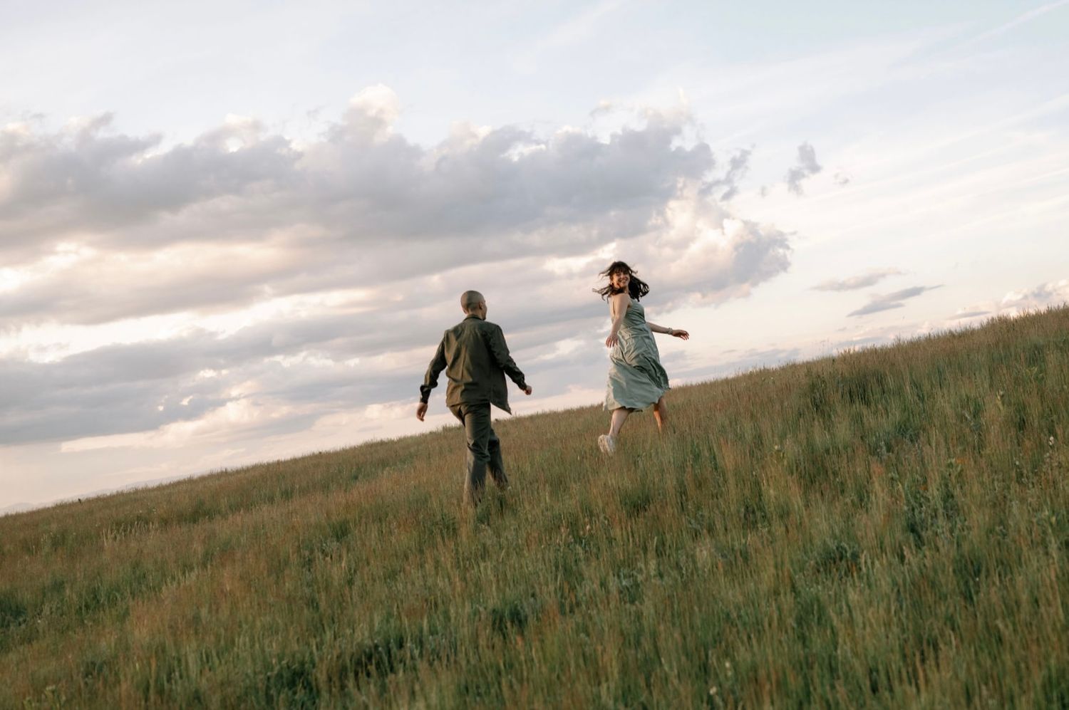 Couple running through a lush green meadow under a dramatic cloudy sky at dusk.