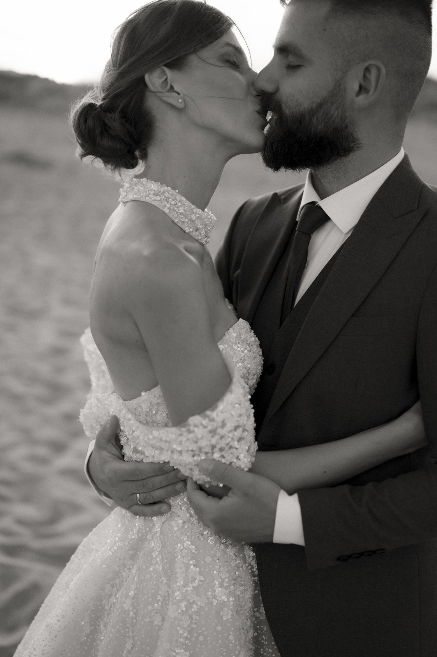 Black and white photo of a couple kissing on a beach, bride in sequined gown, groom in dark suit.