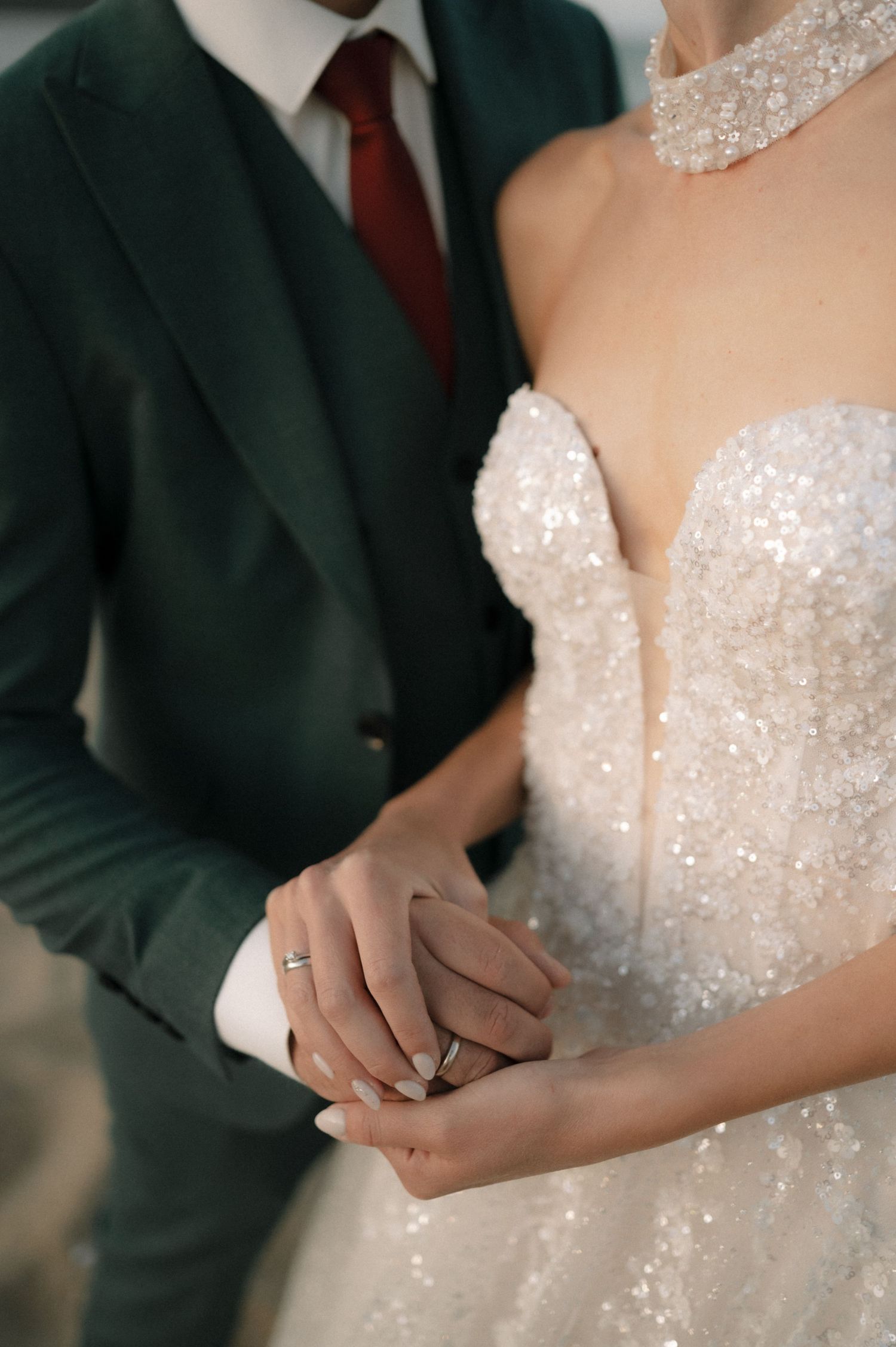 Bride in sequined strapless gown holds hands with groom in dark green suit and red tie at elegant wedding ceremony.