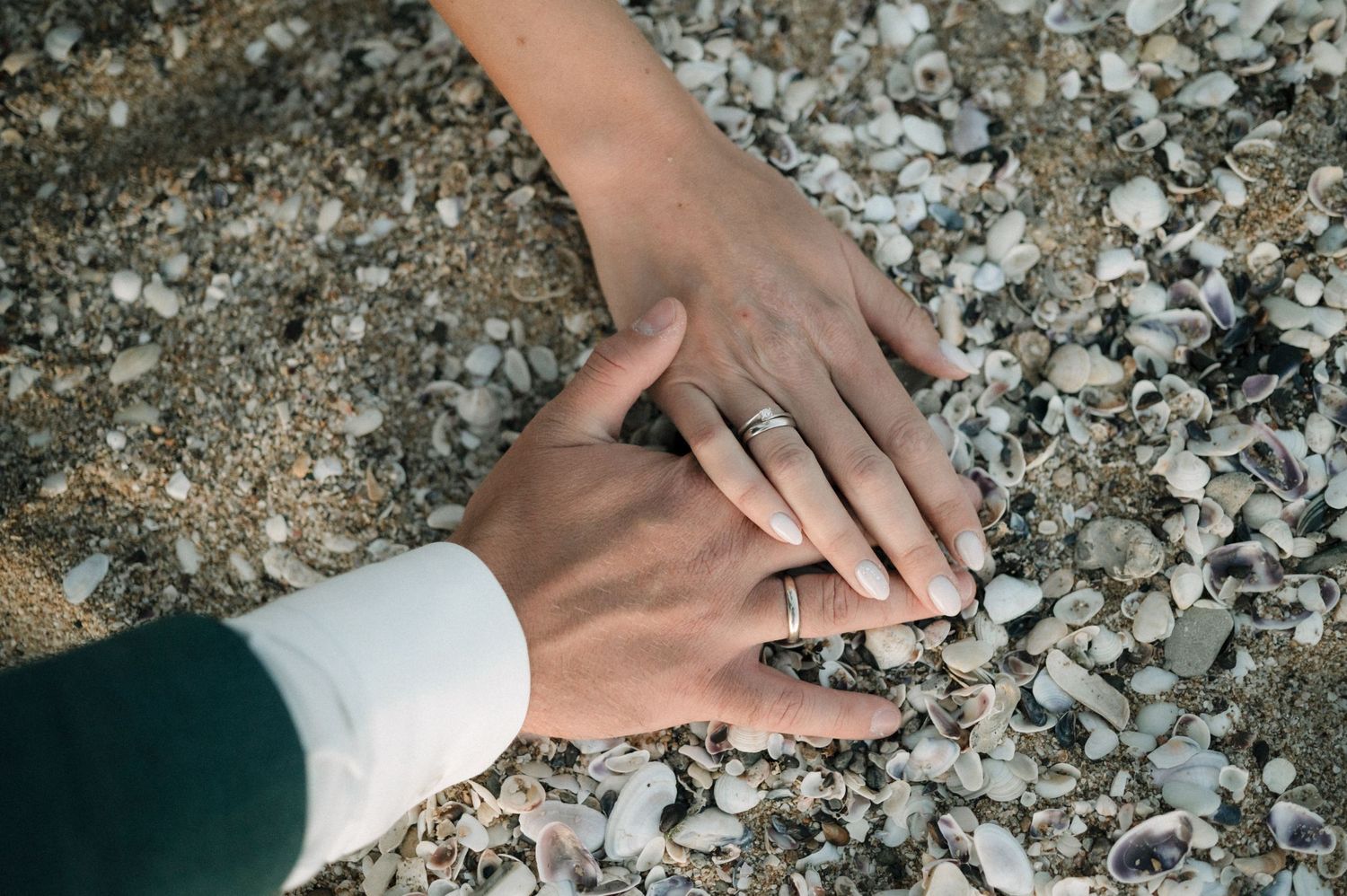 Close-up of bride and groom hands resting on shells and sand, both wearing wedding rings.