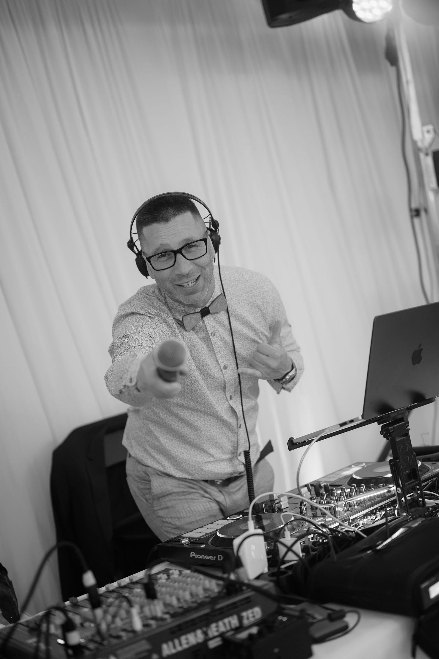 Black and white photo of a smiling DJ wearing headphones and glasses, pointing at camera behind Pioneer DJ equipment.
