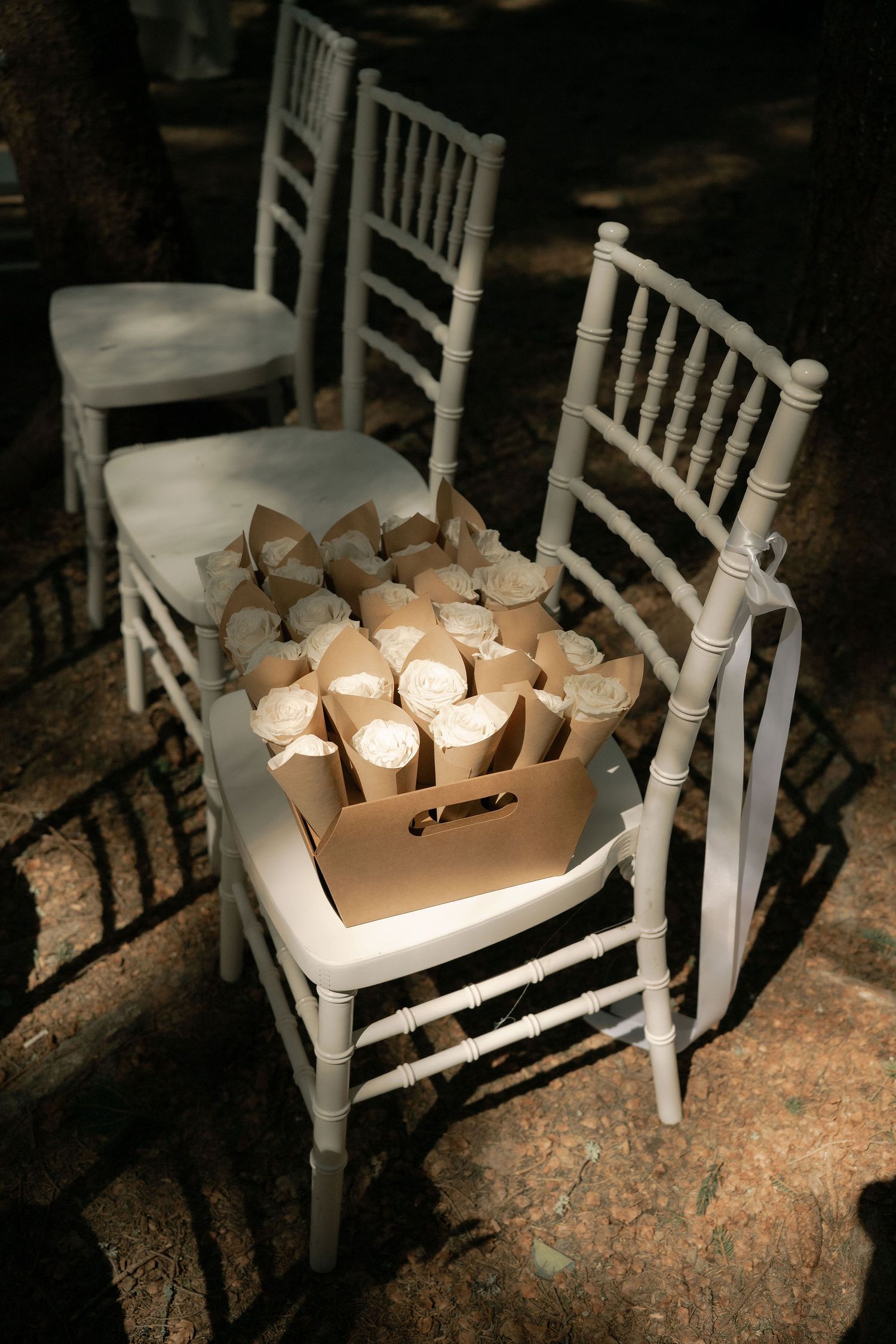 White Chiavari chairs with a cardboard box of paper cone favors for a wedding ceremony outdoors.