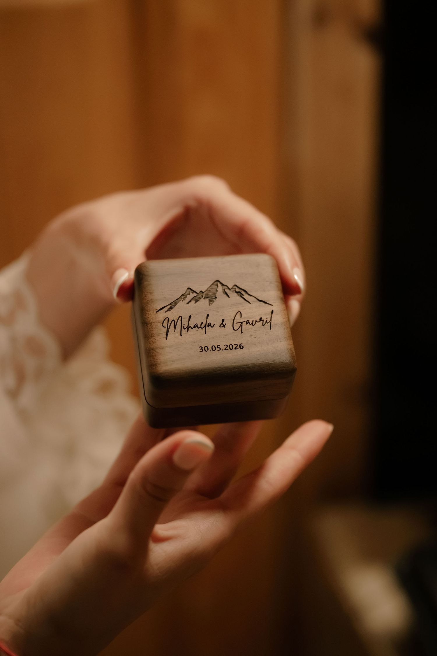 Hands holding a personalized wooden ring box engraved with mountains and names Mihaela & Gavriel, dated 30.05.2026.