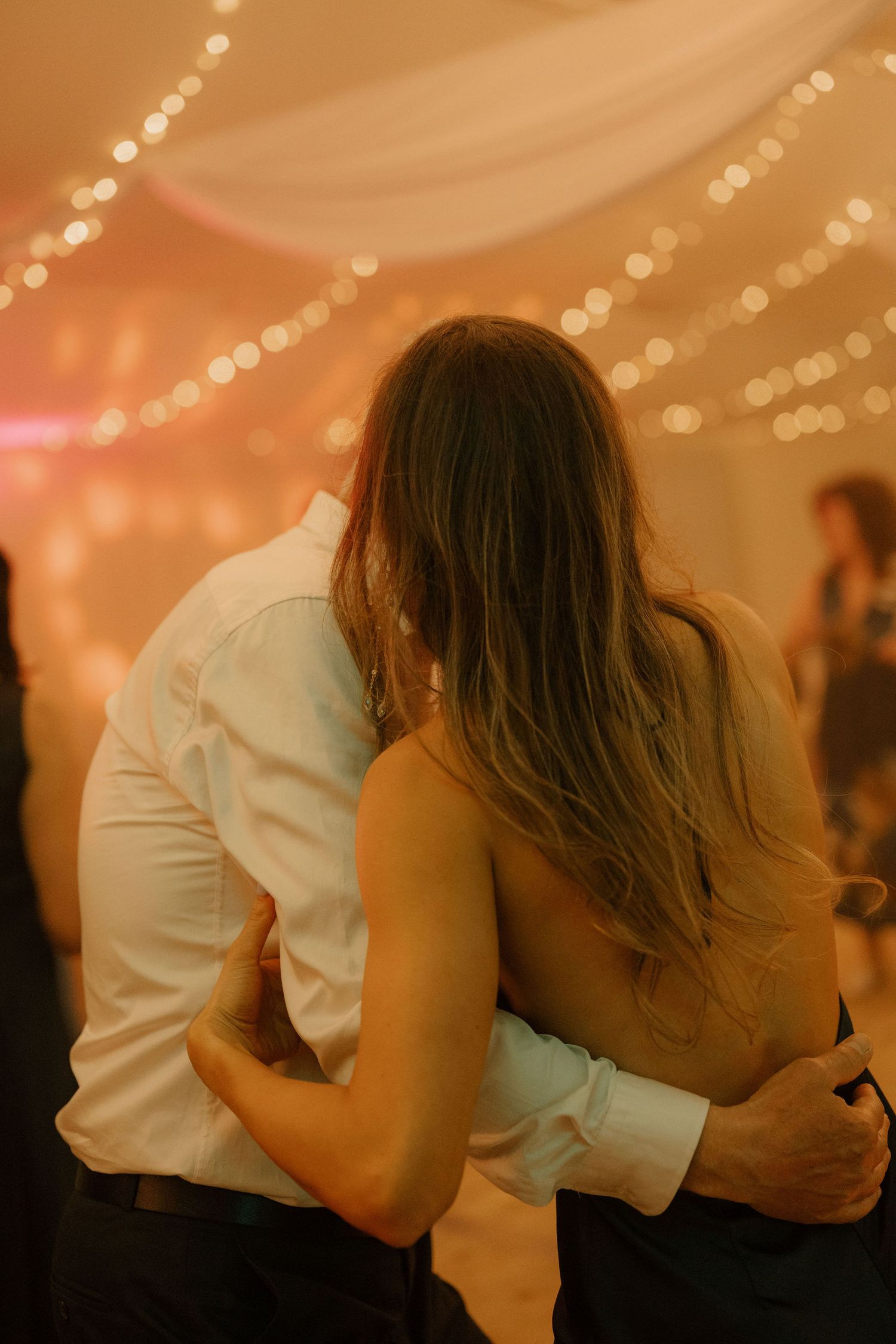 Couple sharing a romantic slow dance embrace at a warmly lit wedding reception with fairy lights.