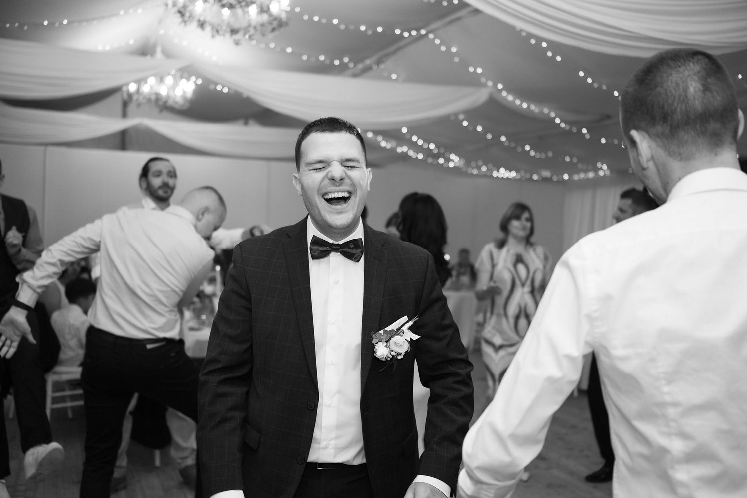 Black and white photo of a laughing groom in a tuxedo dancing joyfully with guests at a wedding reception.