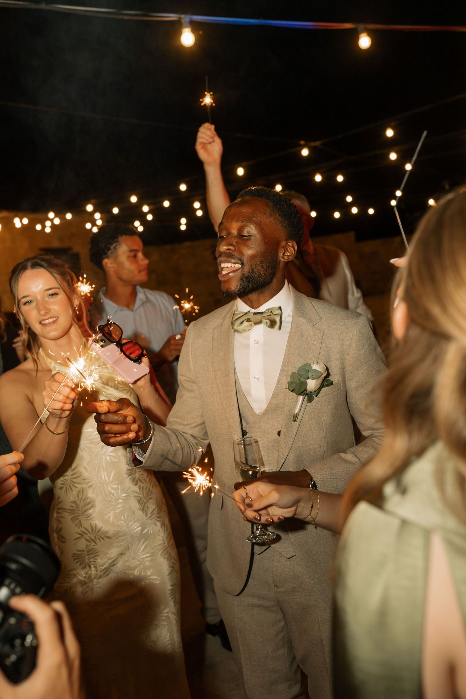 Joyful groom in tan suit with bow tie celebrates with sparklers at outdoor wedding reception with string lights.