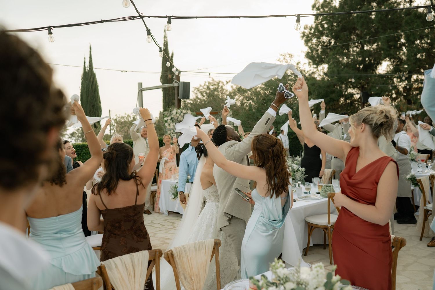 Guests raise glasses in a toast at an outdoor wedding reception with string lights and floral decorations.