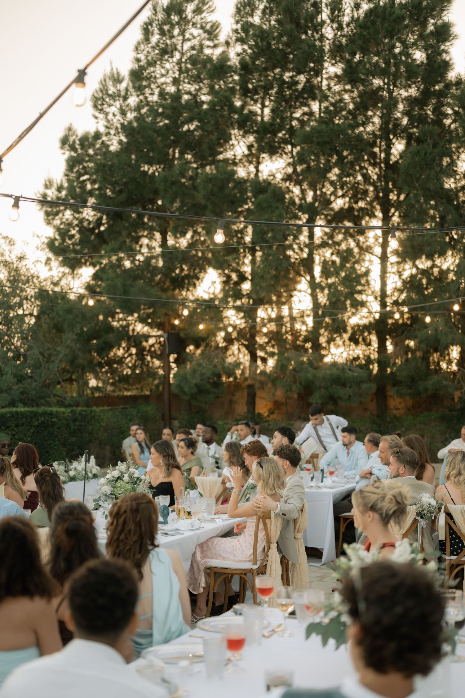 Outdoor wedding reception at sunset with string lights, guests at decorated tables, surrounded by tall pine trees.