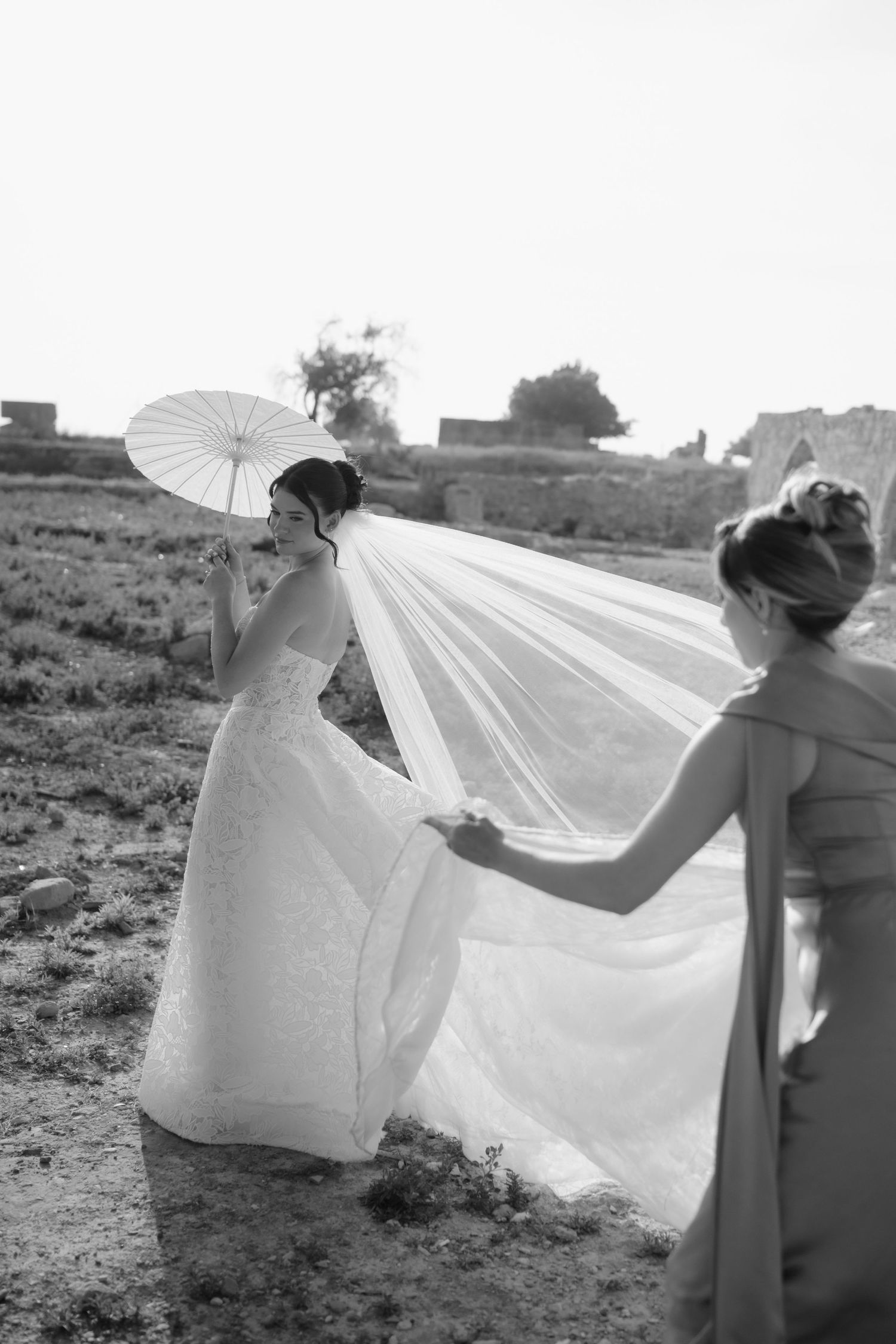 Black and white photo of a bride holding a parasol while a bridesmaid arranges her flowing veil outdoors.