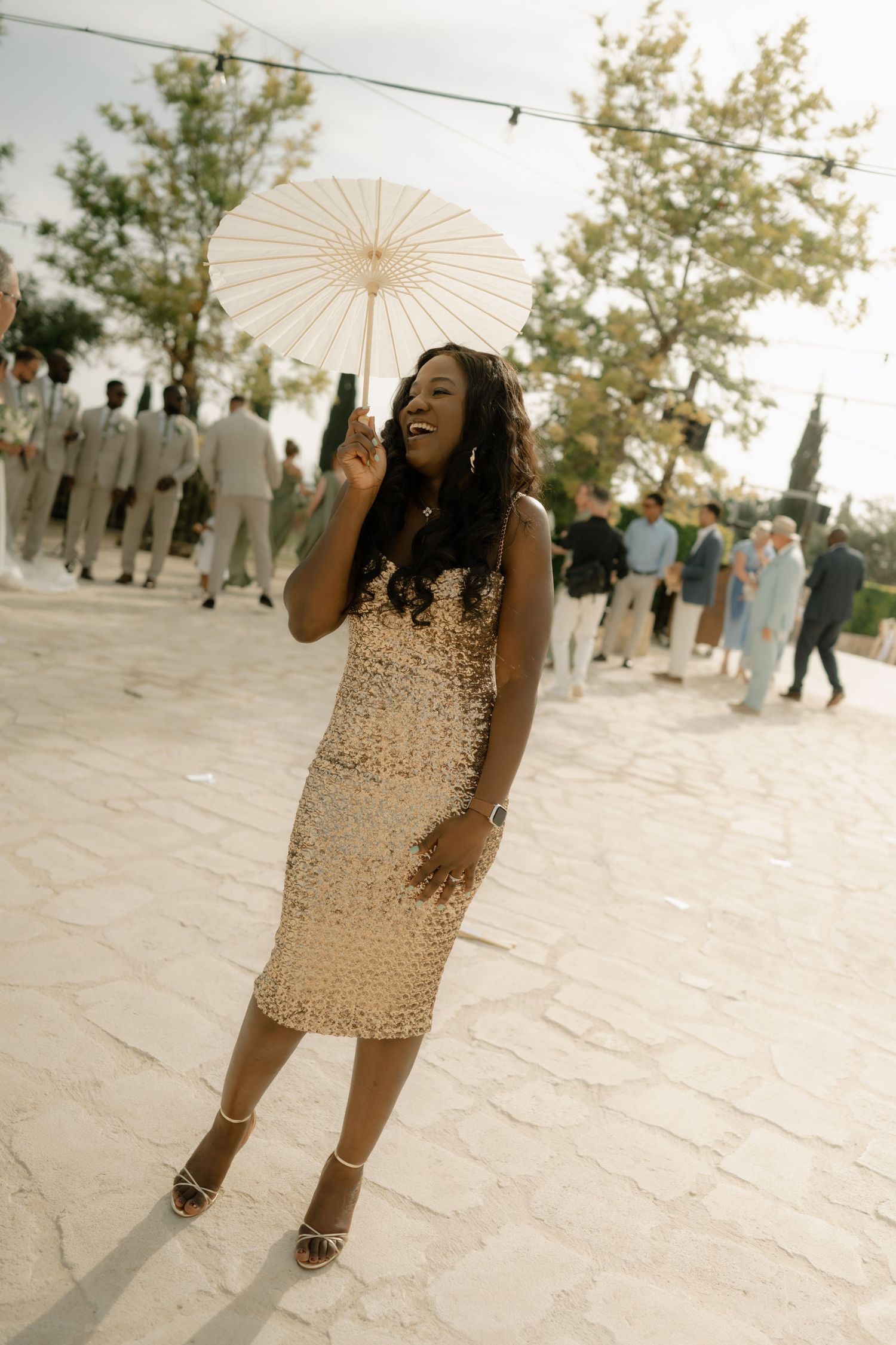 Elegant woman in gold sequin dress holding white parasol at outdoor event on stone patio.