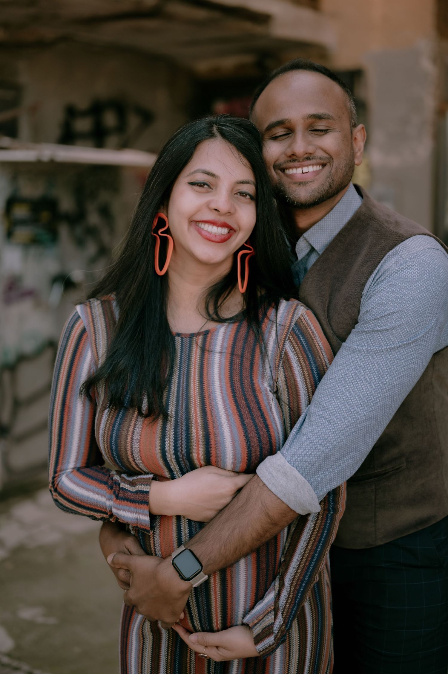 Happy couple embracing outdoors, pregnant woman in striped dress with red earrings, man in vest smiling warmly.