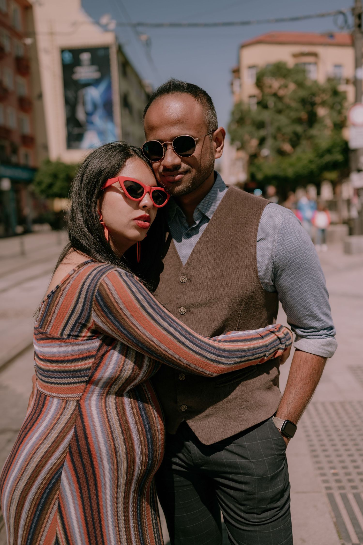 Stylish couple posing on a sunny urban street, woman in striped dress with red sunglasses, man in vest and shades.