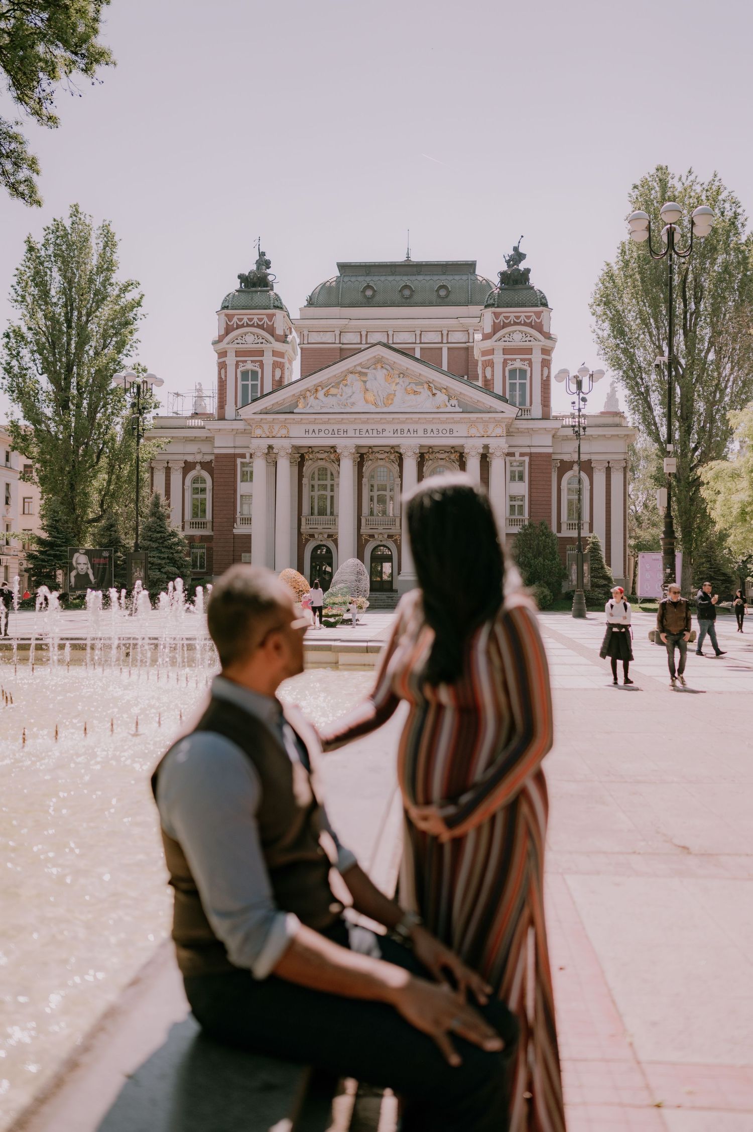 Couple posing in front of a grand historic building with fountains, woman in striped dress touching man's face.