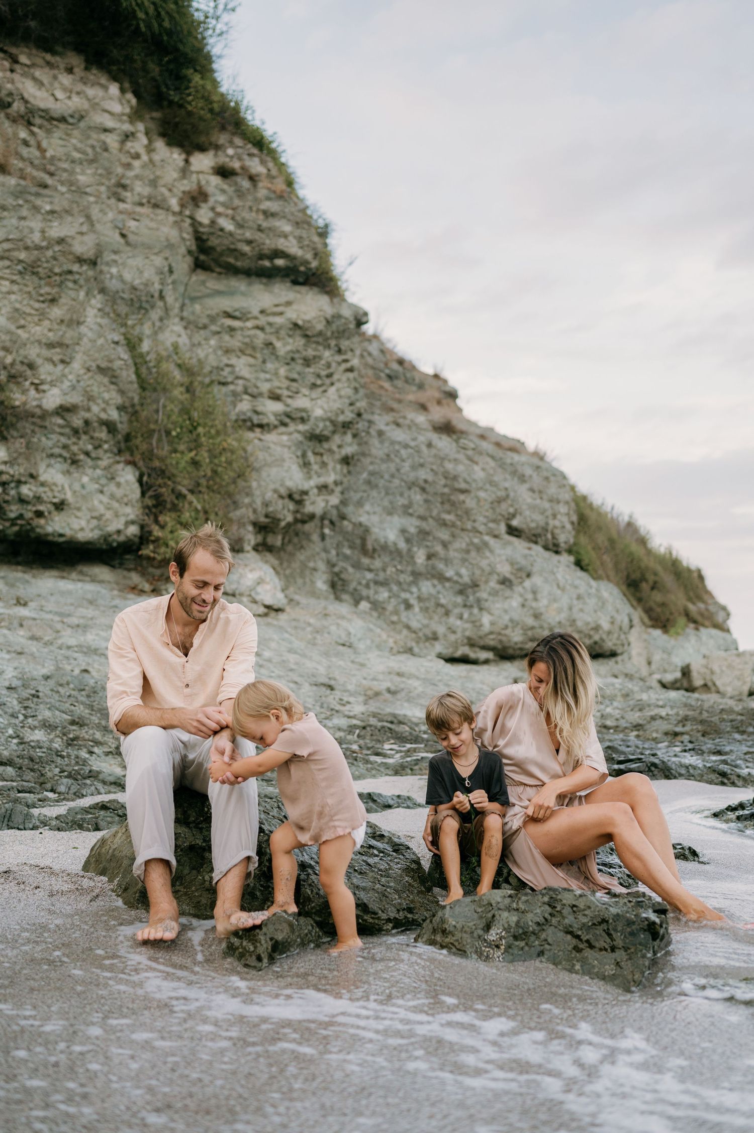 Family of four enjoying a relaxing beach day on rocky shore with scenic coastal cliffs in the background.