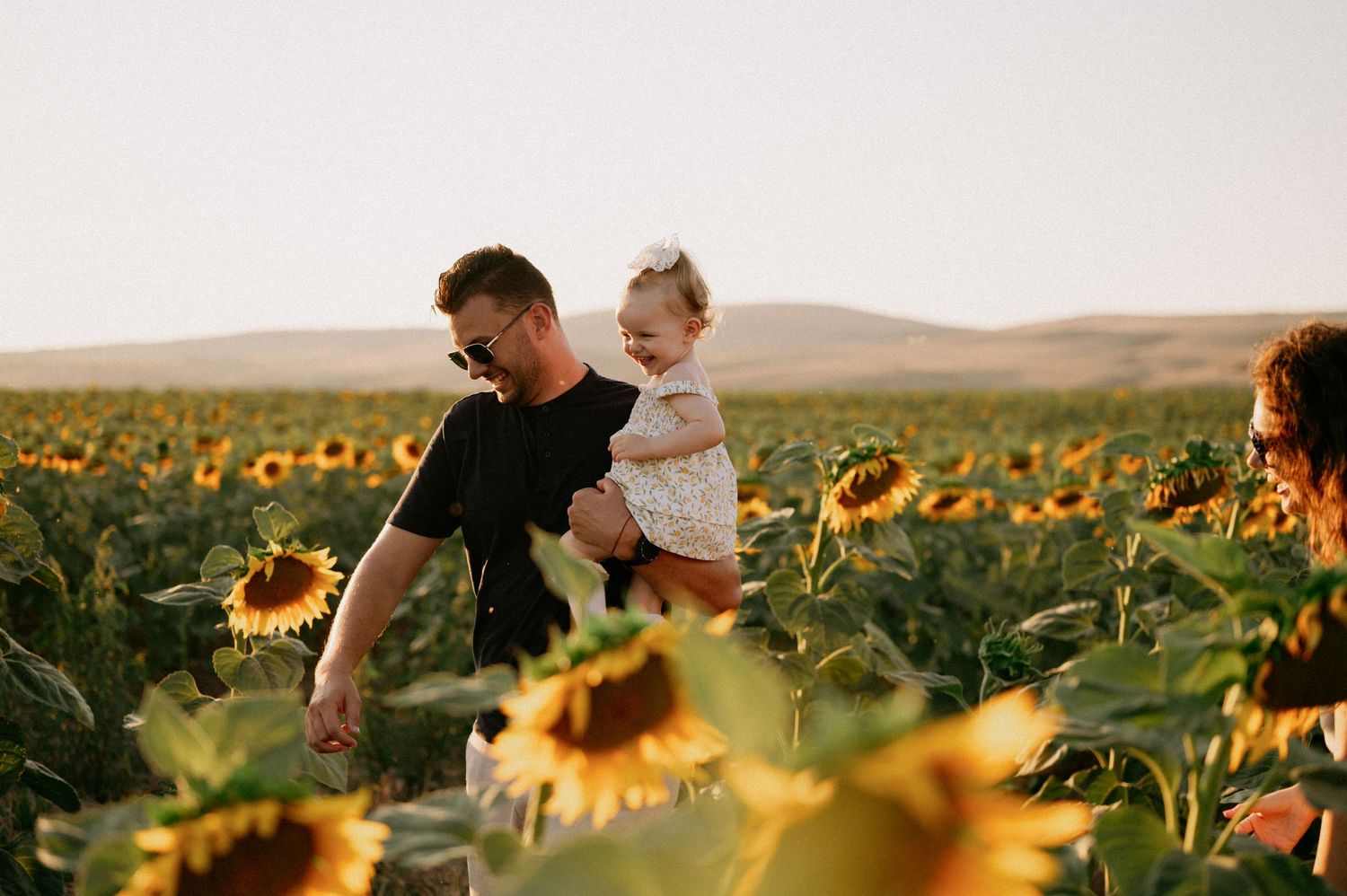 A father holds his toddler in a golden sunflower field while a woman walks beside them on a sunny day.