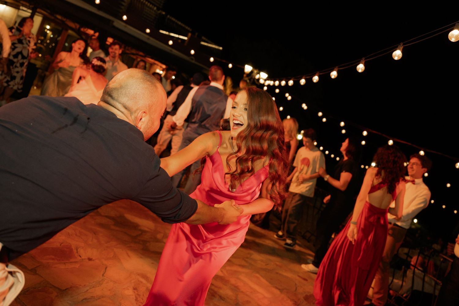 Dancers move across a dimly lit dance floor under glowing string lights at an evening event.