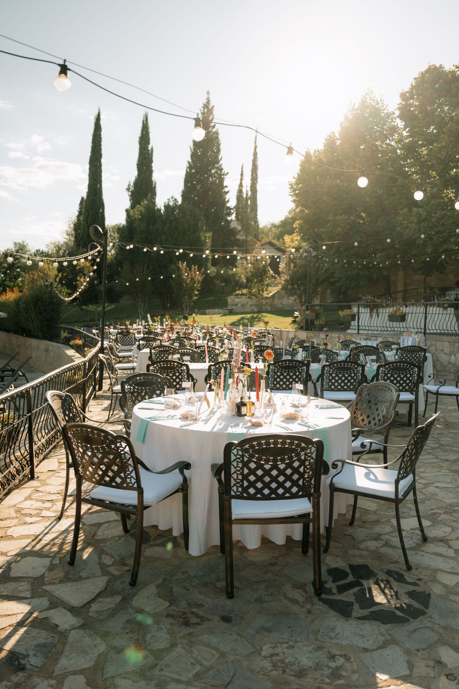 Outdoor wedding reception setup with string lights and white tablecloths on a stone patio surrounded by cypress trees at sunset.