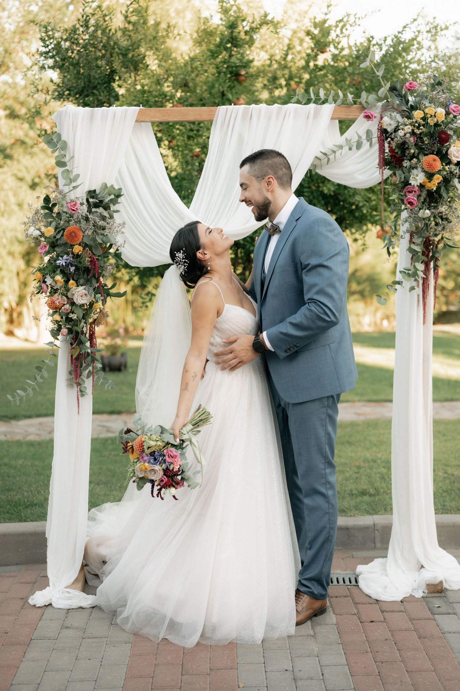 A couple shares a romantic kiss under an elegant white draped wedding arch decorated with pink and burgundy florals.