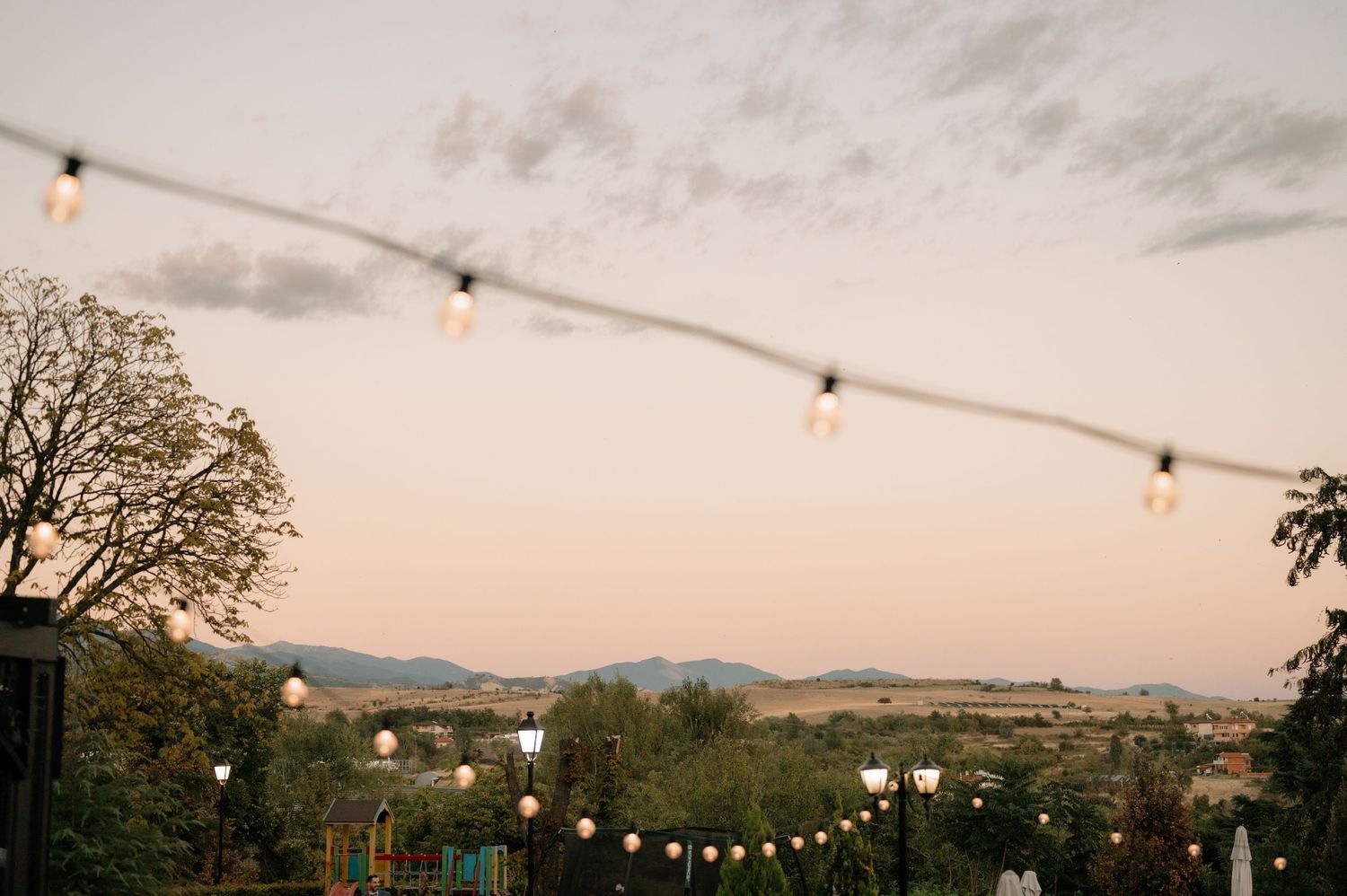String lights hang above an outdoor venue with mountains and trees silhouetted against a dusky sky at sunset.