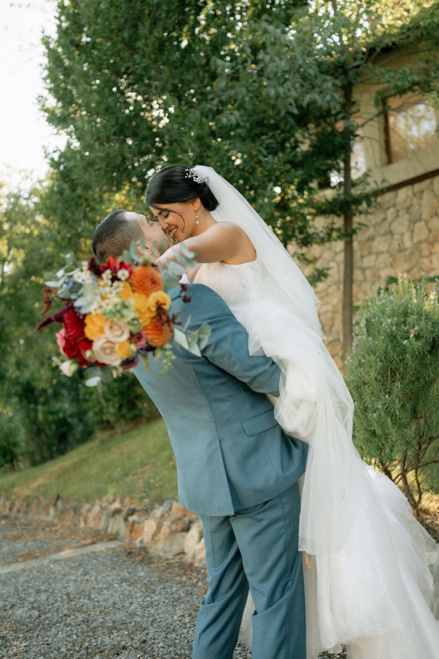 A couple in wedding attire embrace and dance together on a gravel path surrounded by lush greenery at sunset.