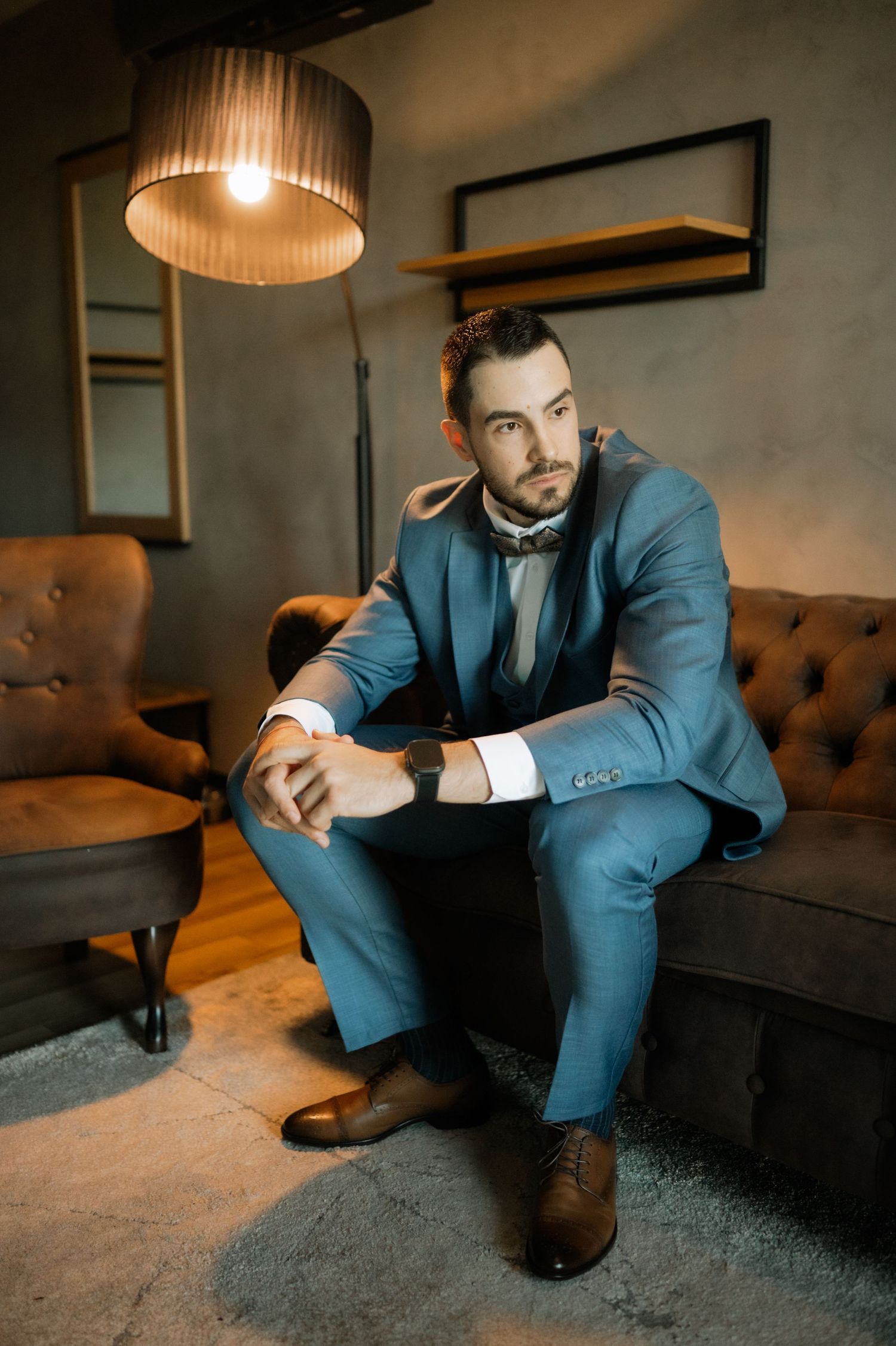 A person in a blue suit sits thoughtfully on a gray couch in a dimly lit room with pendant lighting.