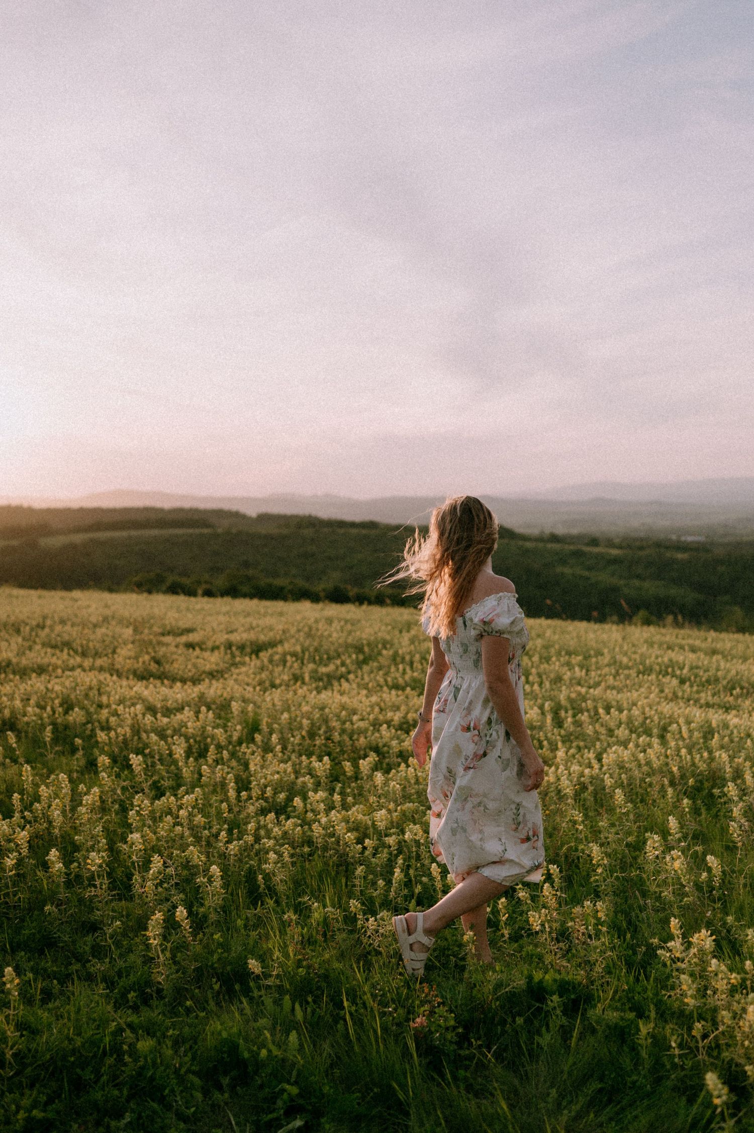 Woman in a floral dress walking through a field of yellow wildflowers at sunset.