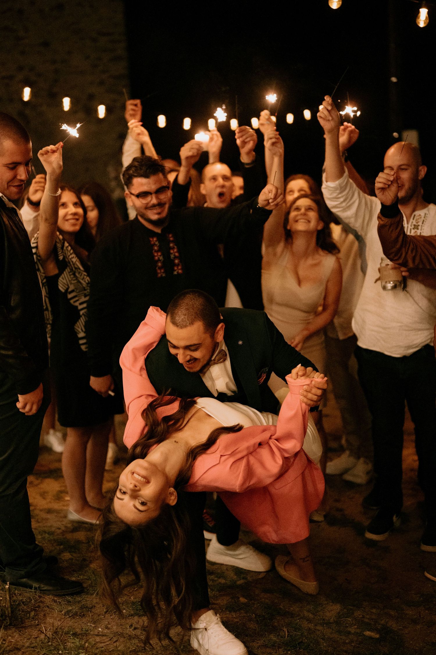 Energetic dance moves captured during an outdoor nighttime celebration with string lights.
