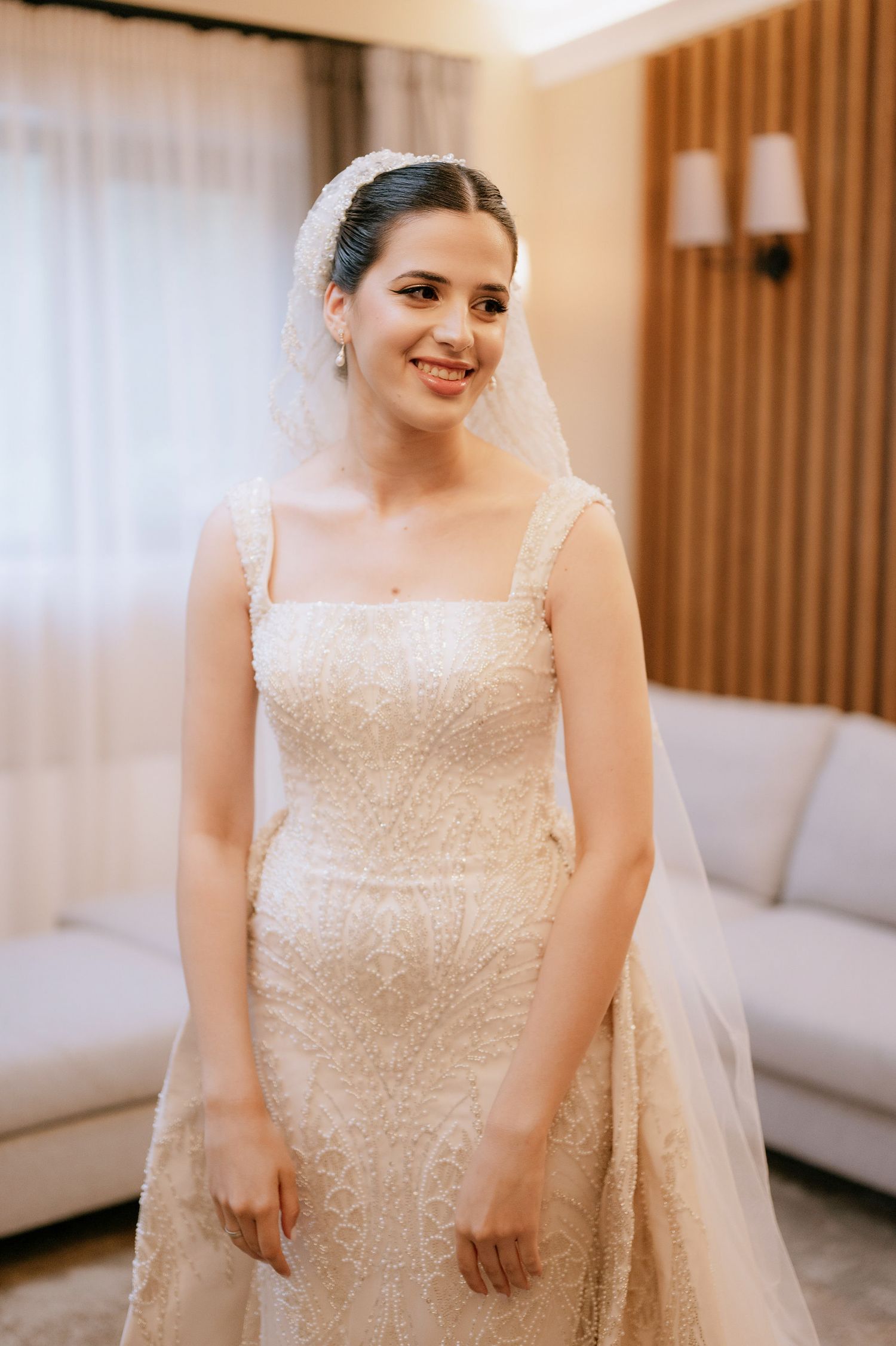 A bride wearing a stunning white beaded wedding gown with square neckline and delicate veil in an elegant room.