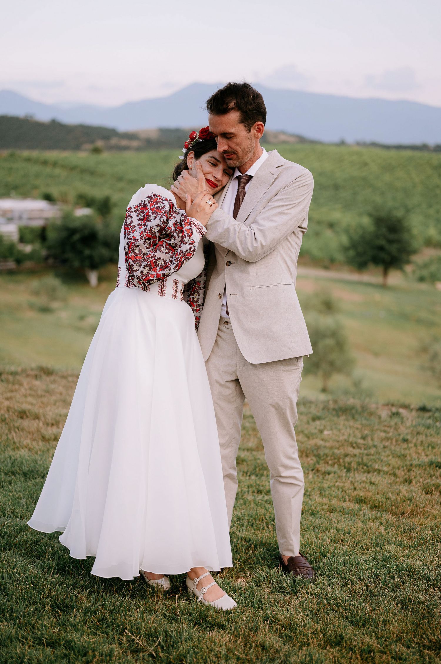 Couple in wedding attire shares intimate dance on green hillside with mountains in background.