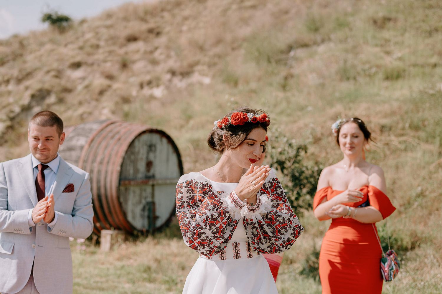 Wedding party members in light suits and floral dresses celebrate on a grassy hillside.