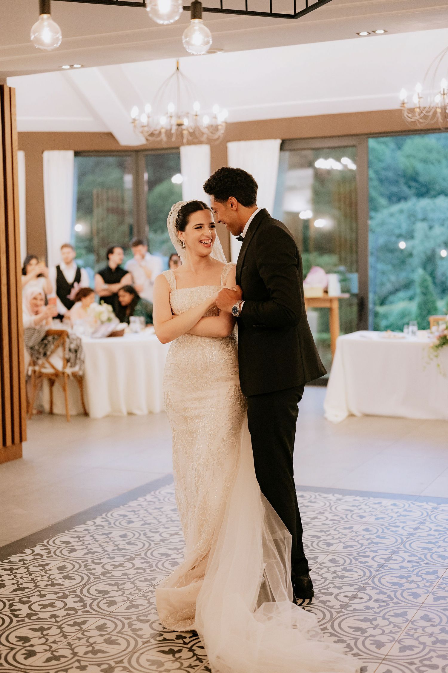 A romantic first dance in an elegant reception hall with chandelier lighting and floor-length windows.