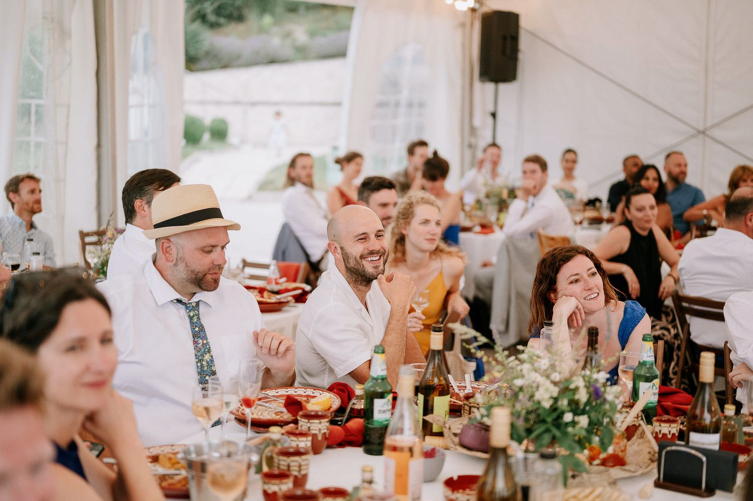 Wedding reception guests dining at decorated tables with floral centerpieces and natural lighting.