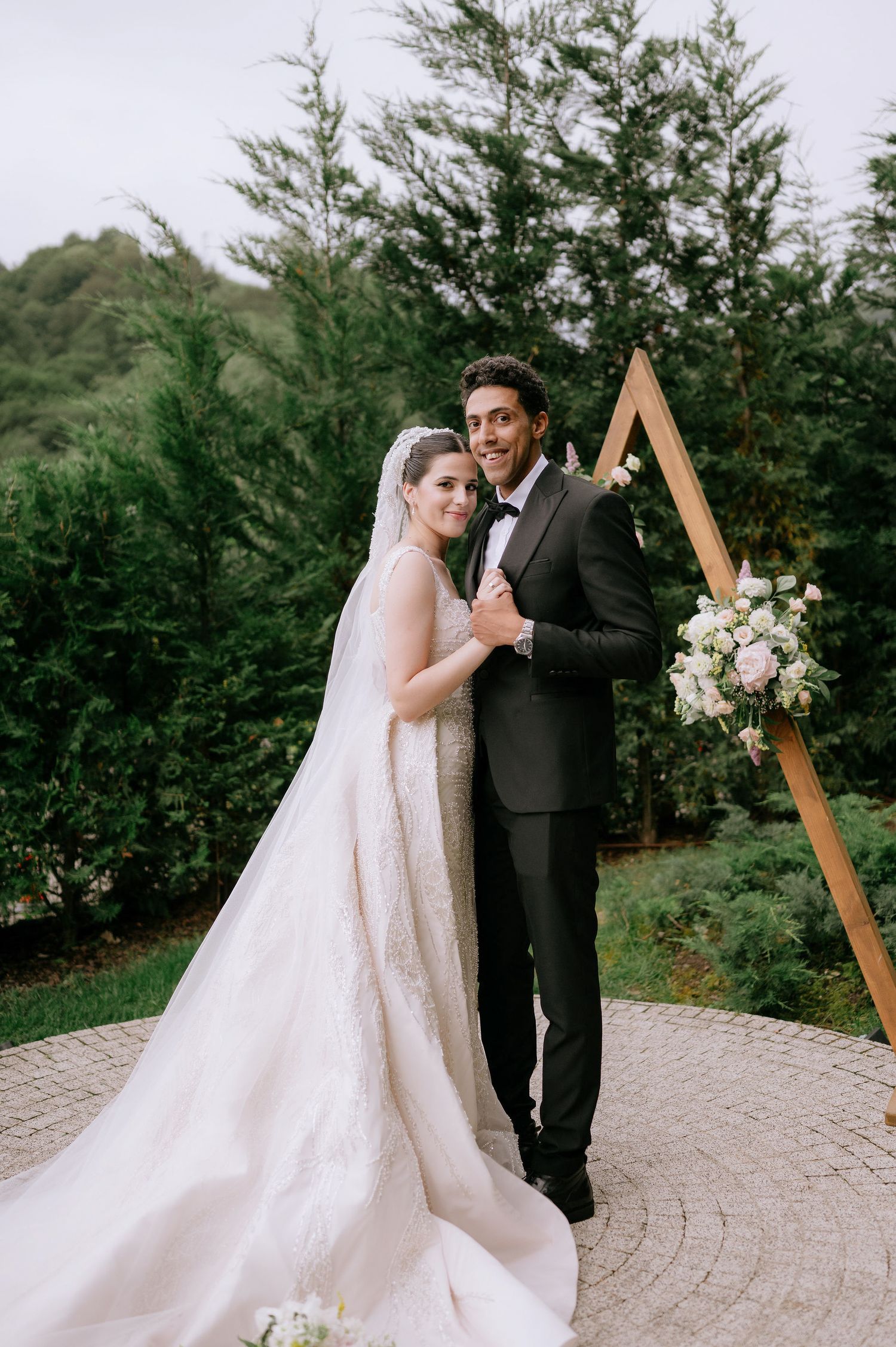 Wedding couple posing in front of a wooden triangle arch decorated with white flowers.
