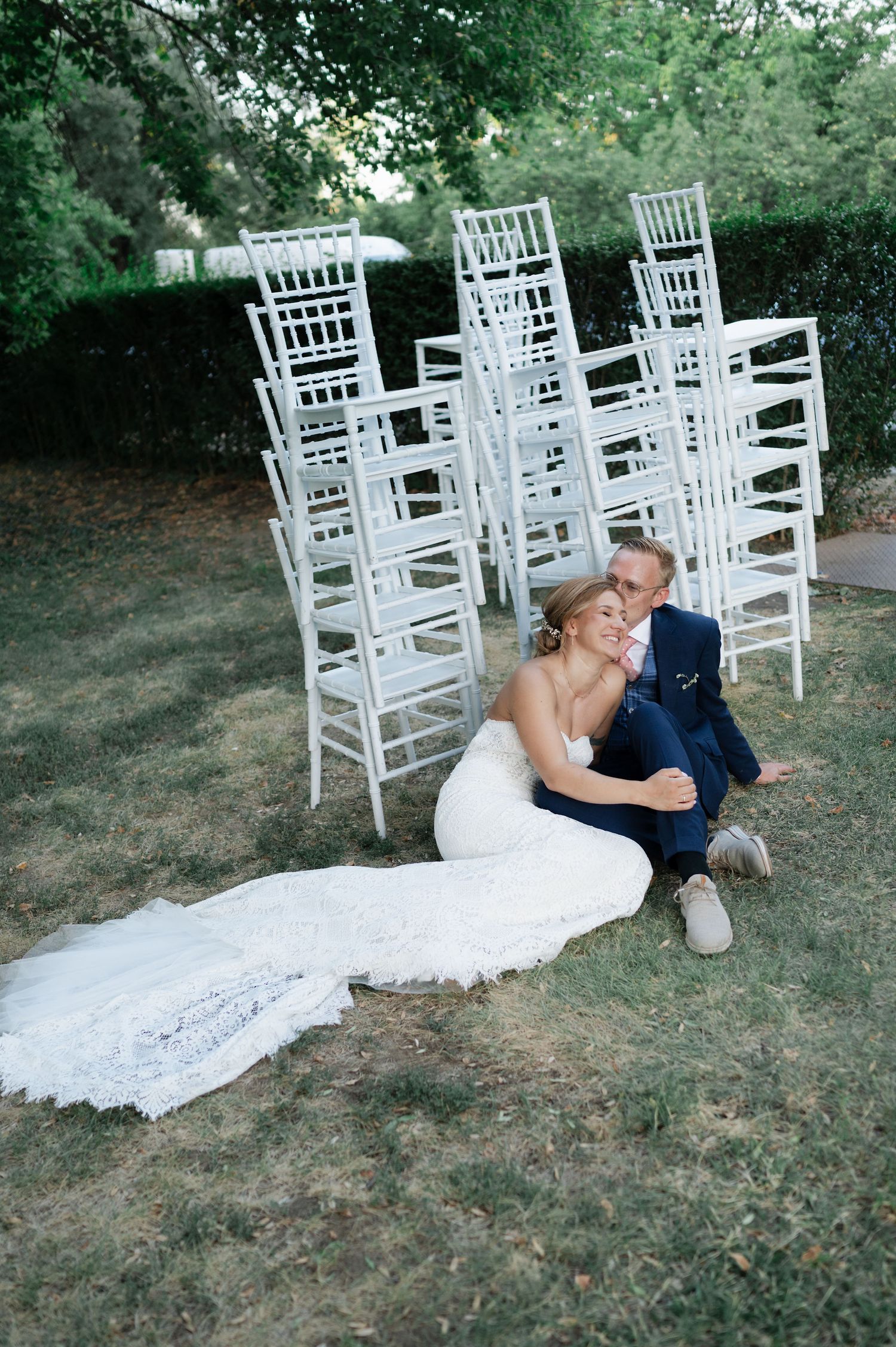 Wedding couple shares joyful moment while sitting on grass near white ceremony chairs.