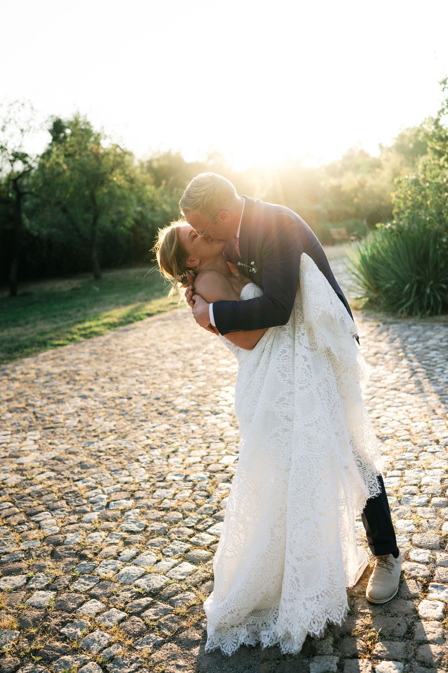 A couple in wedding attire embraces on a sunlit cobblestone pathway surrounded by greenery.