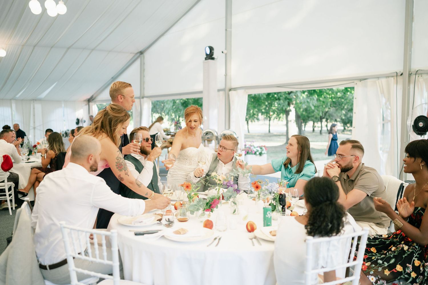 Wide view of wedding reception tent interior with guests seated at decorated round tables.