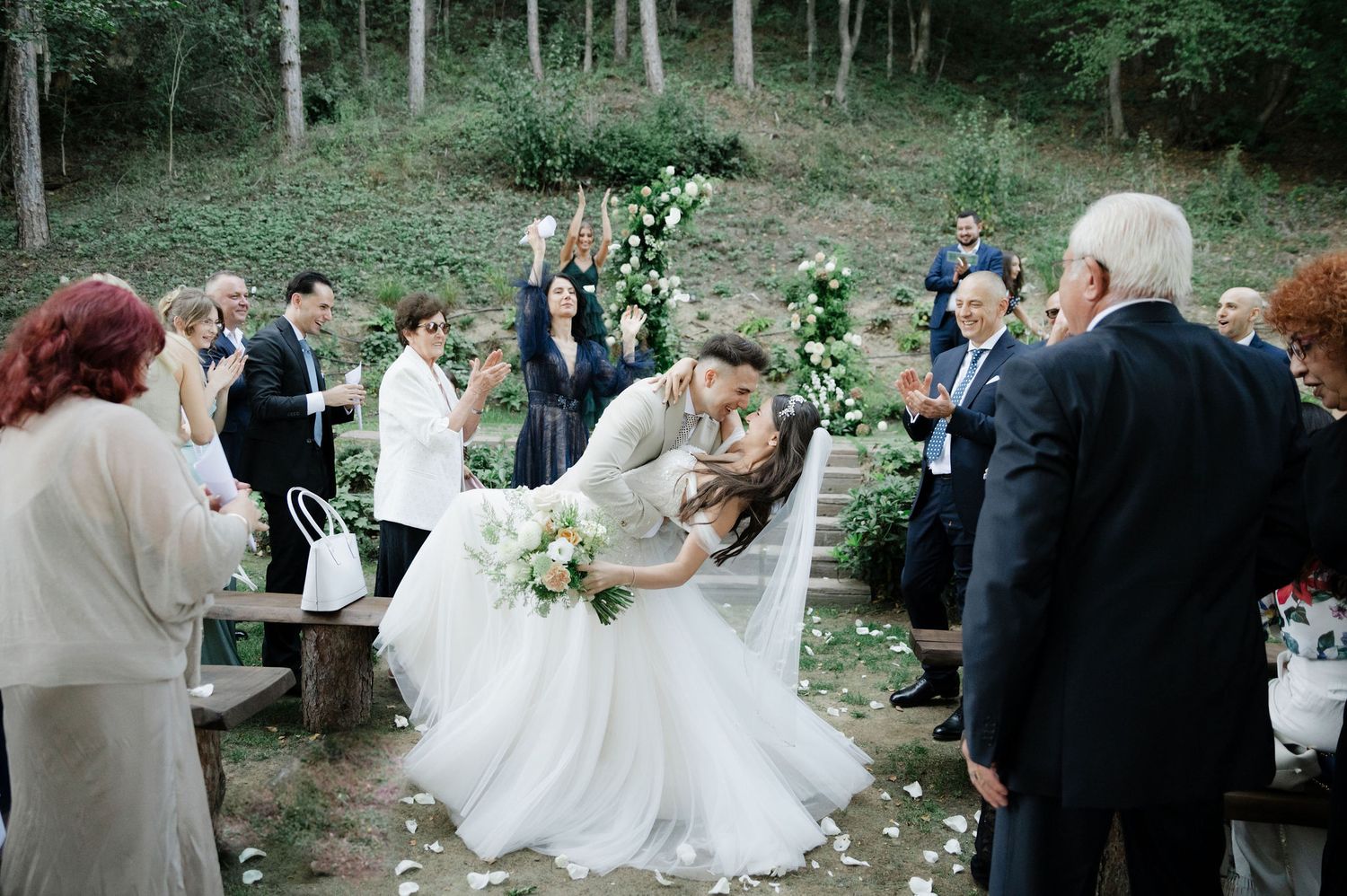 Elegant outdoor wedding ceremony with guests gathered on stone steps in a forest setting.