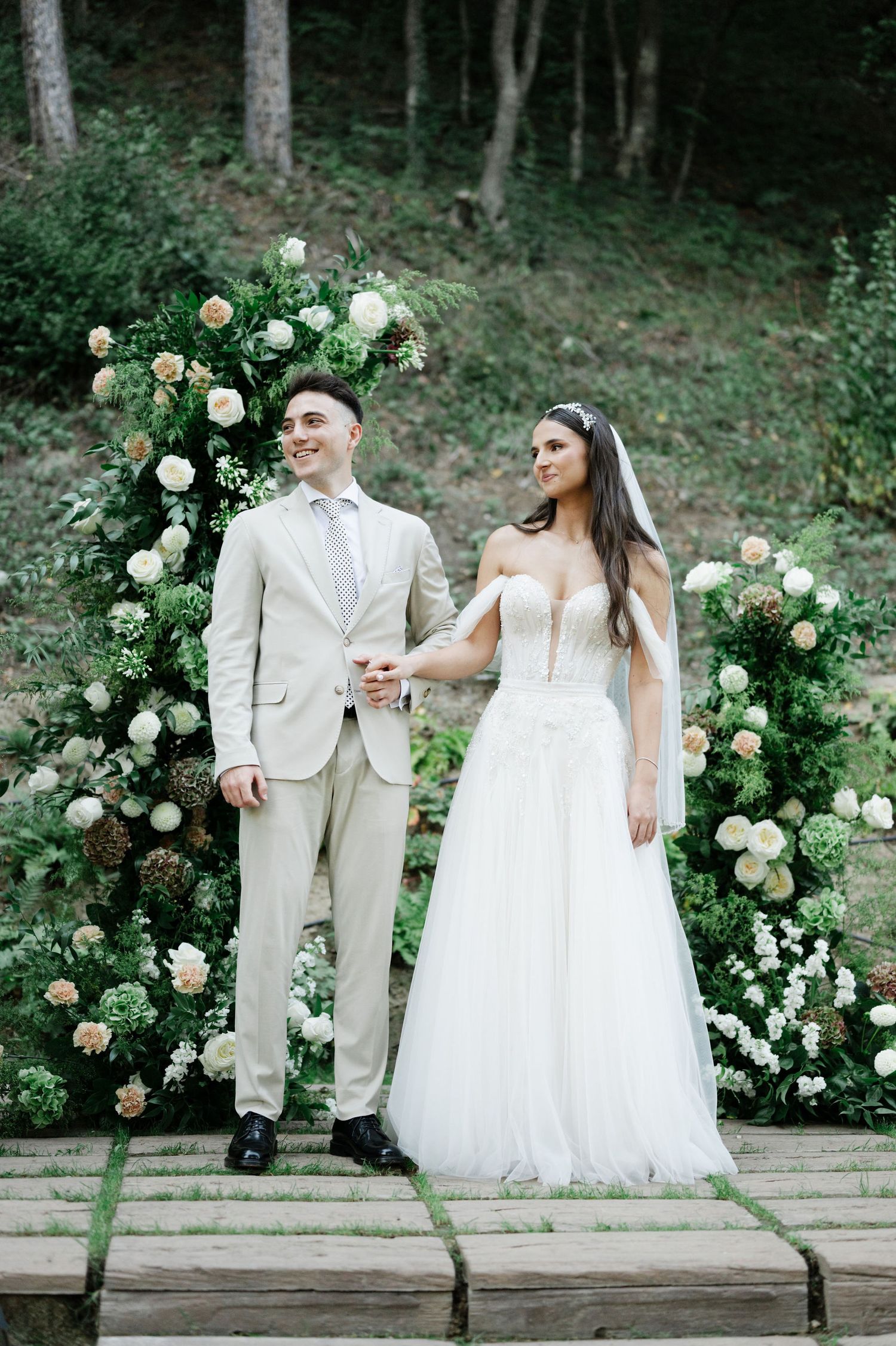 Elegant wedding couple poses on stone platform with lush floral arch in forest setting.