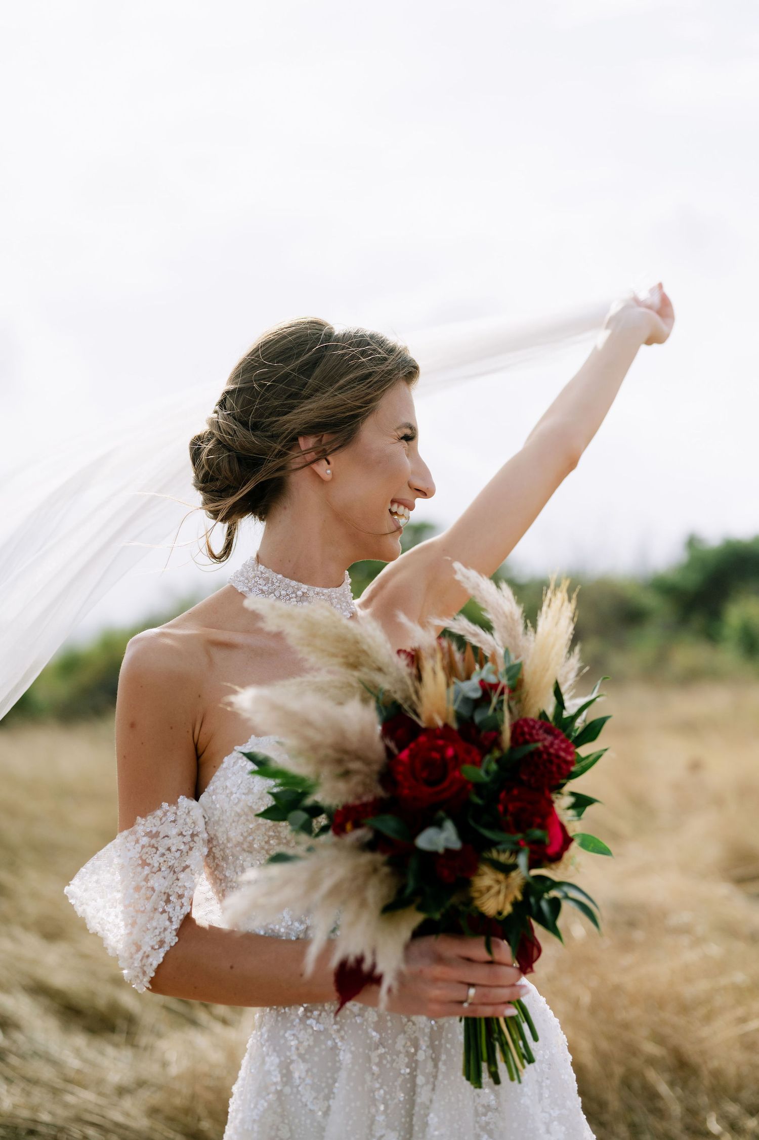A bride in an elegant white dress playfully holds her veil while carrying a vibrant autumn bouquet.