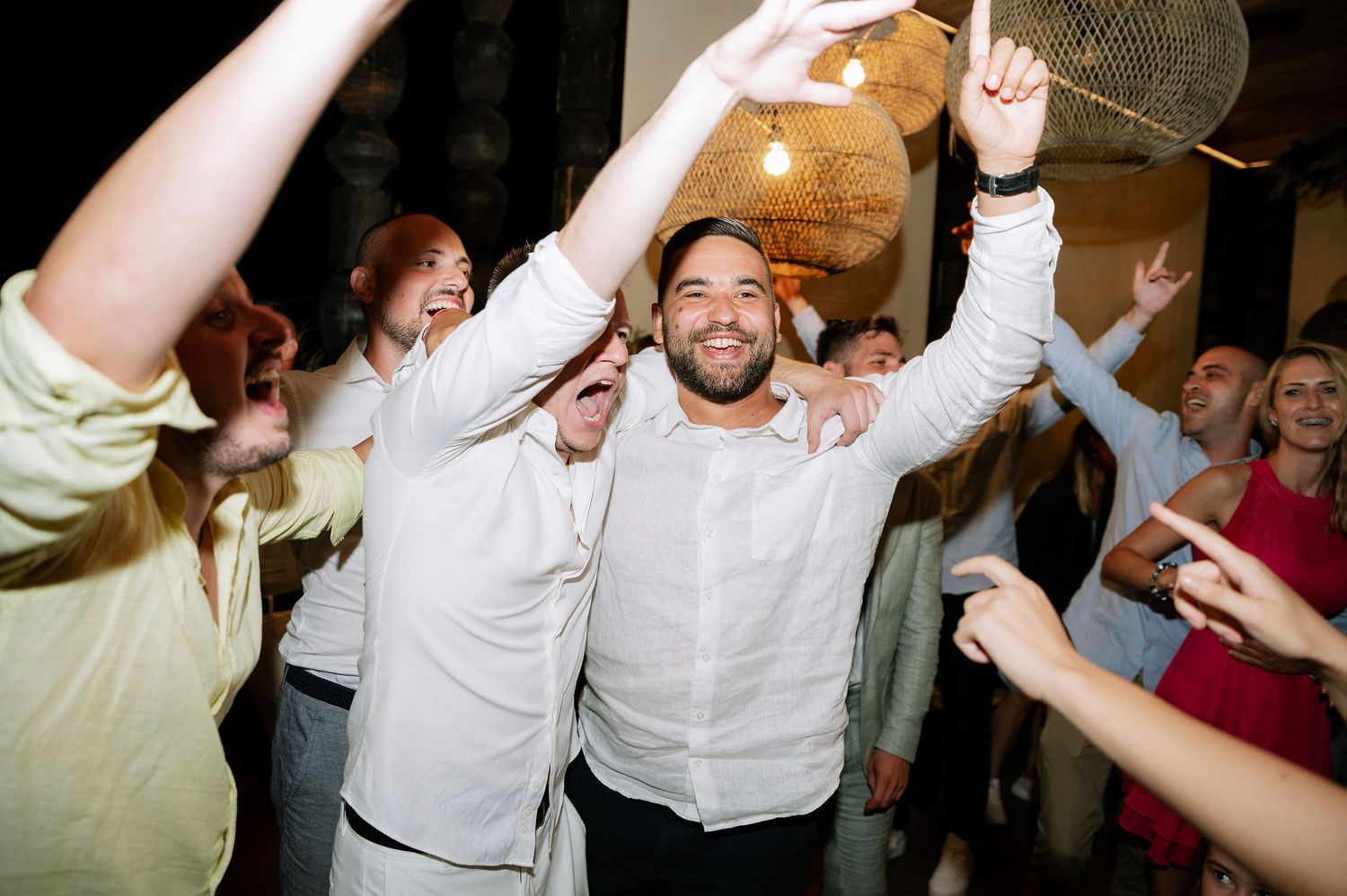 Party guests dancing and celebrating with raised arms under warm indoor lighting.