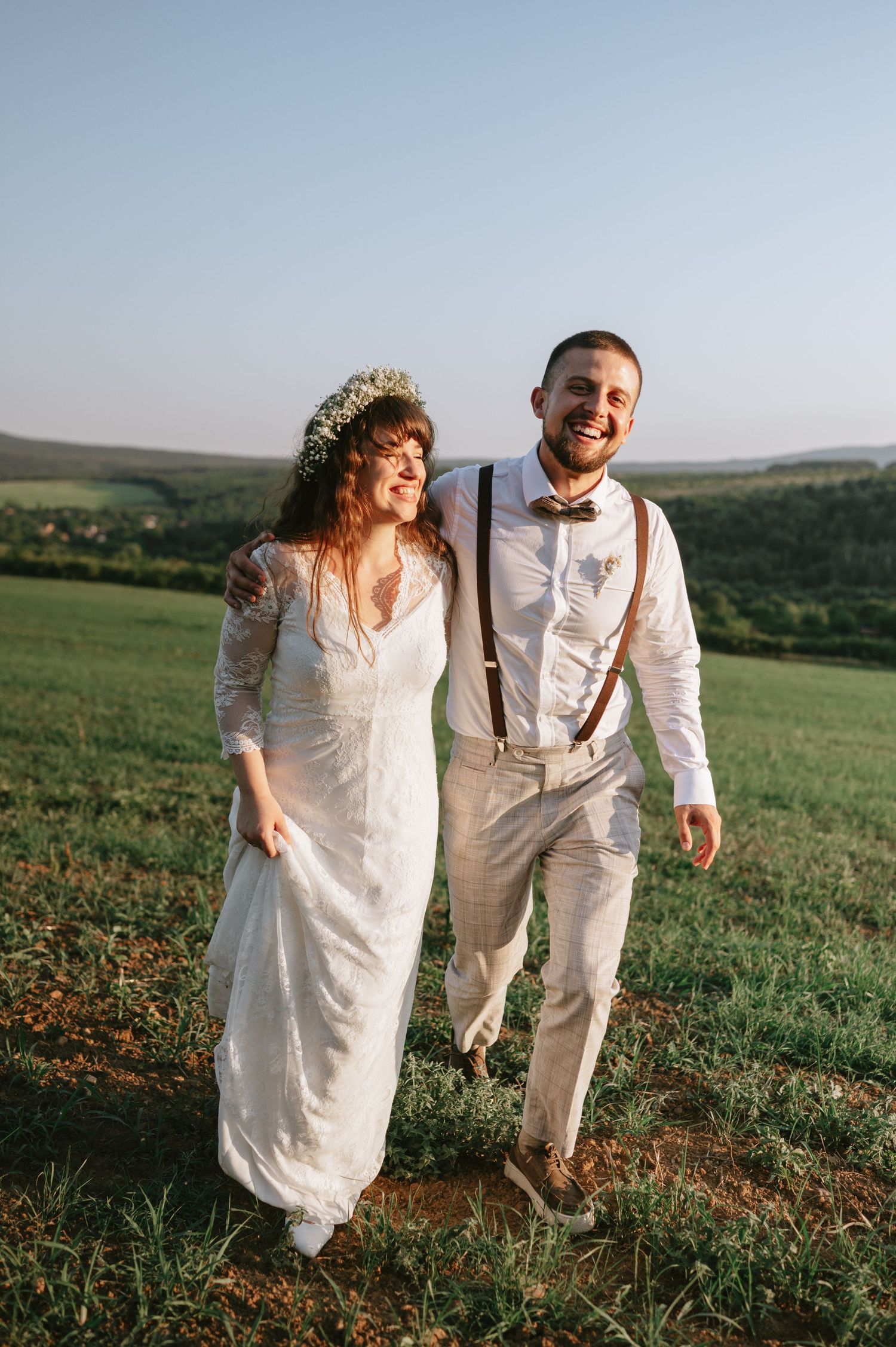 Two people in white wedding attire laugh together while walking on a grassy hillside.