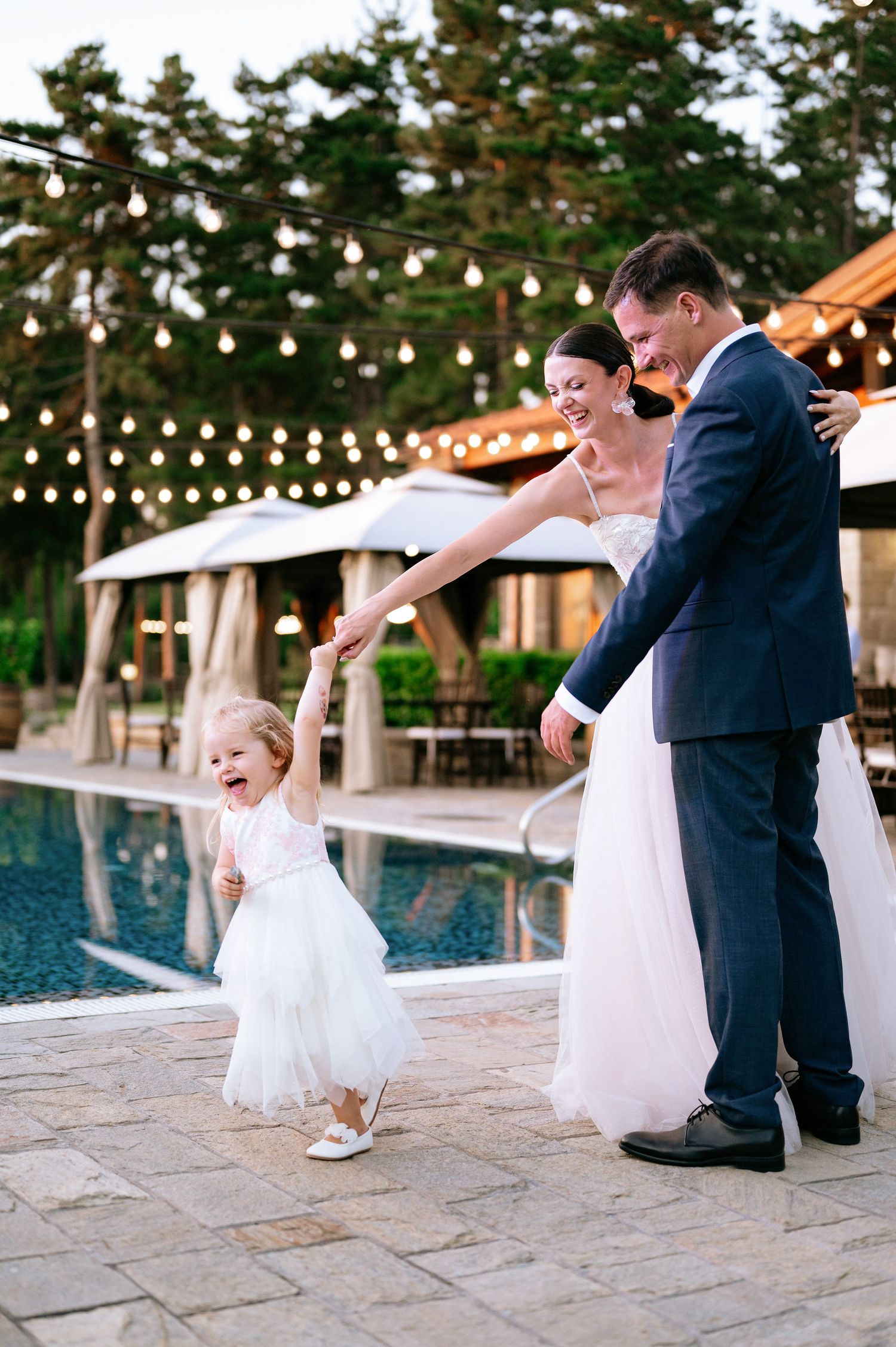 Couple performing playful dance moves on stone patio near pool during wedding reception.