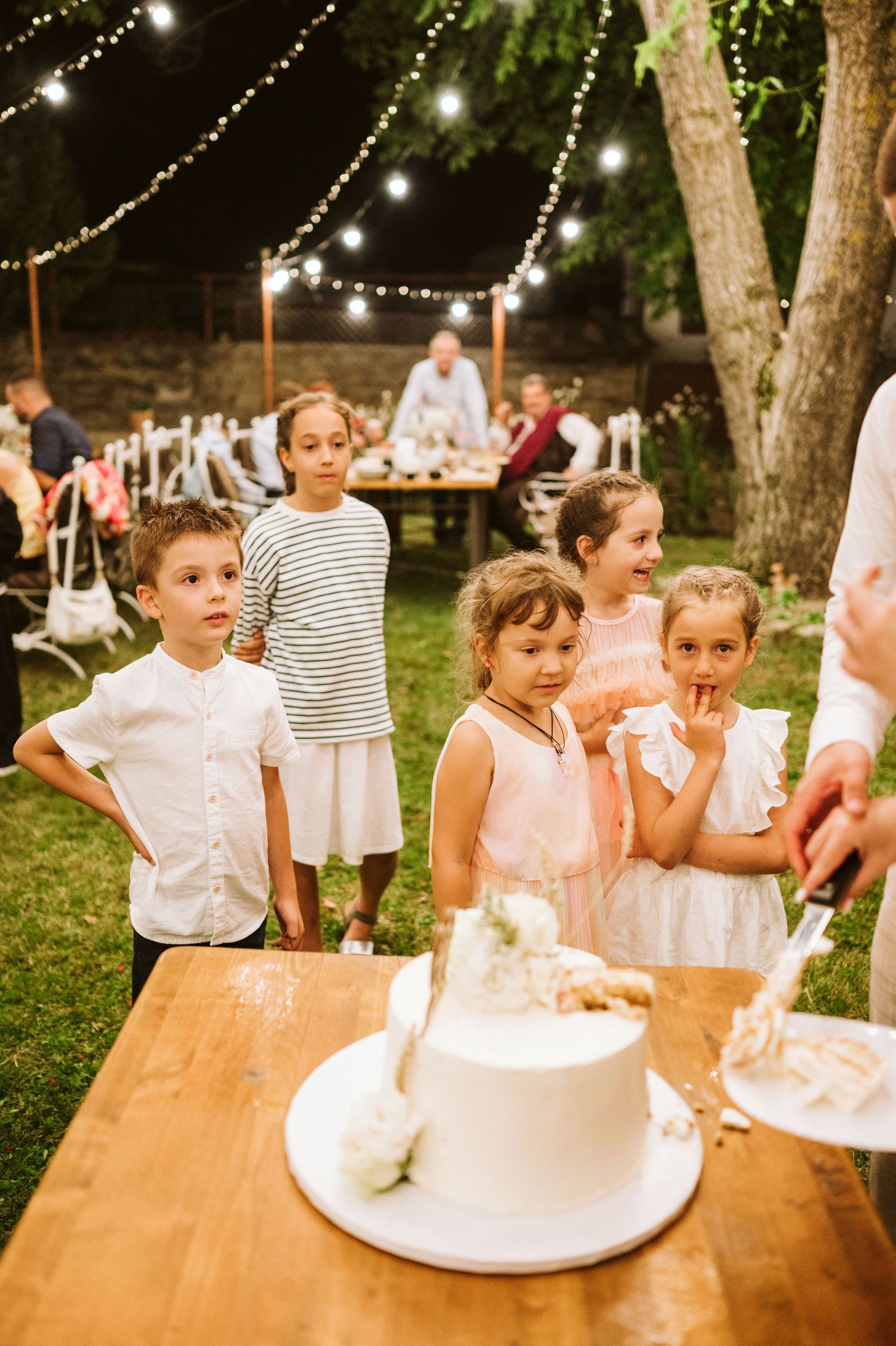 Children gather excitedly around a cake at an outdoor wedding reception with string lights overhead.