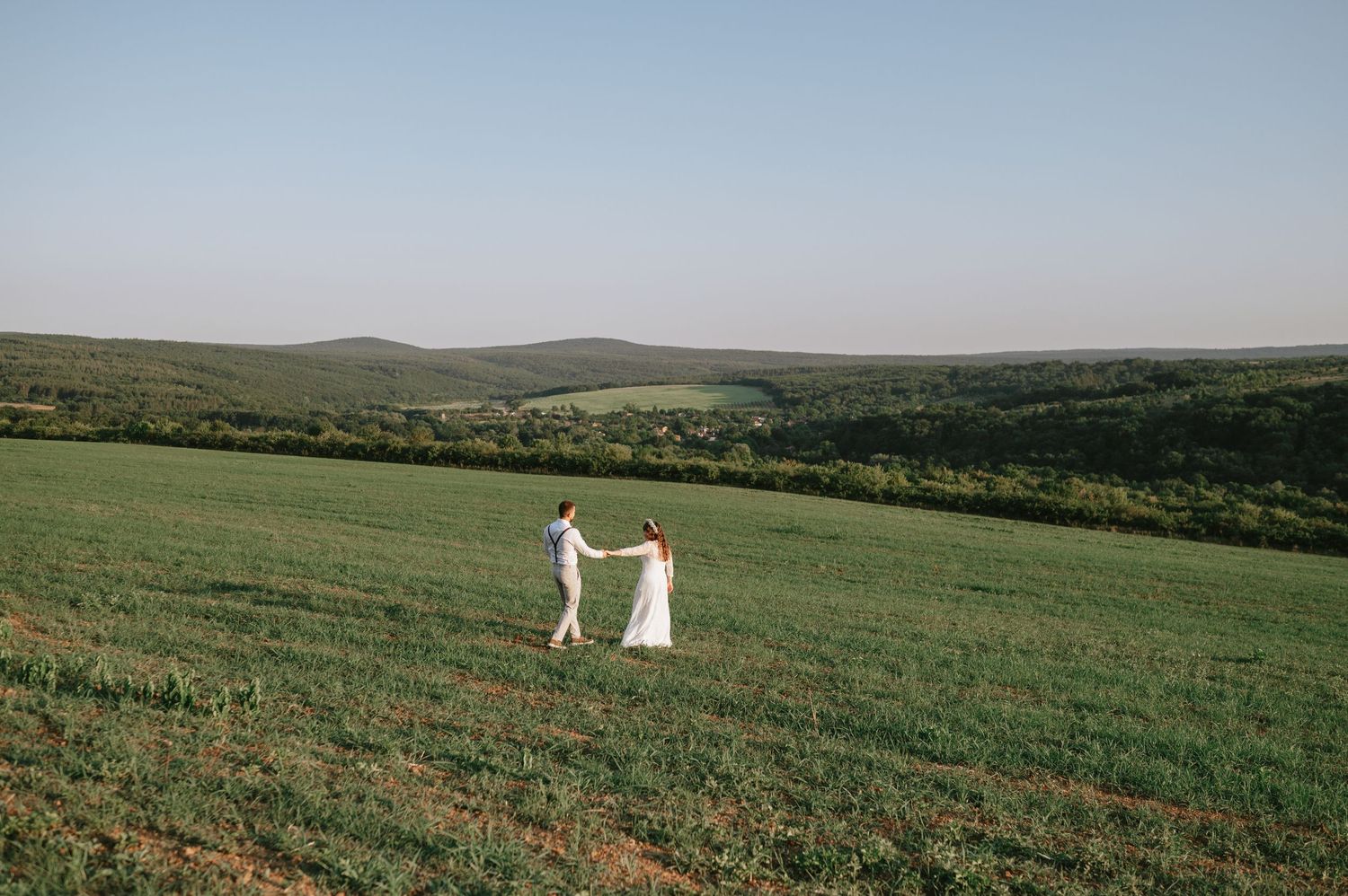 Two people in white stand together in a panoramic hilltop meadow.