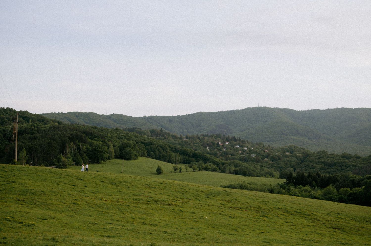 Rolling green hills and mountains stretch across a scenic mountain landscape under cloudy skies.