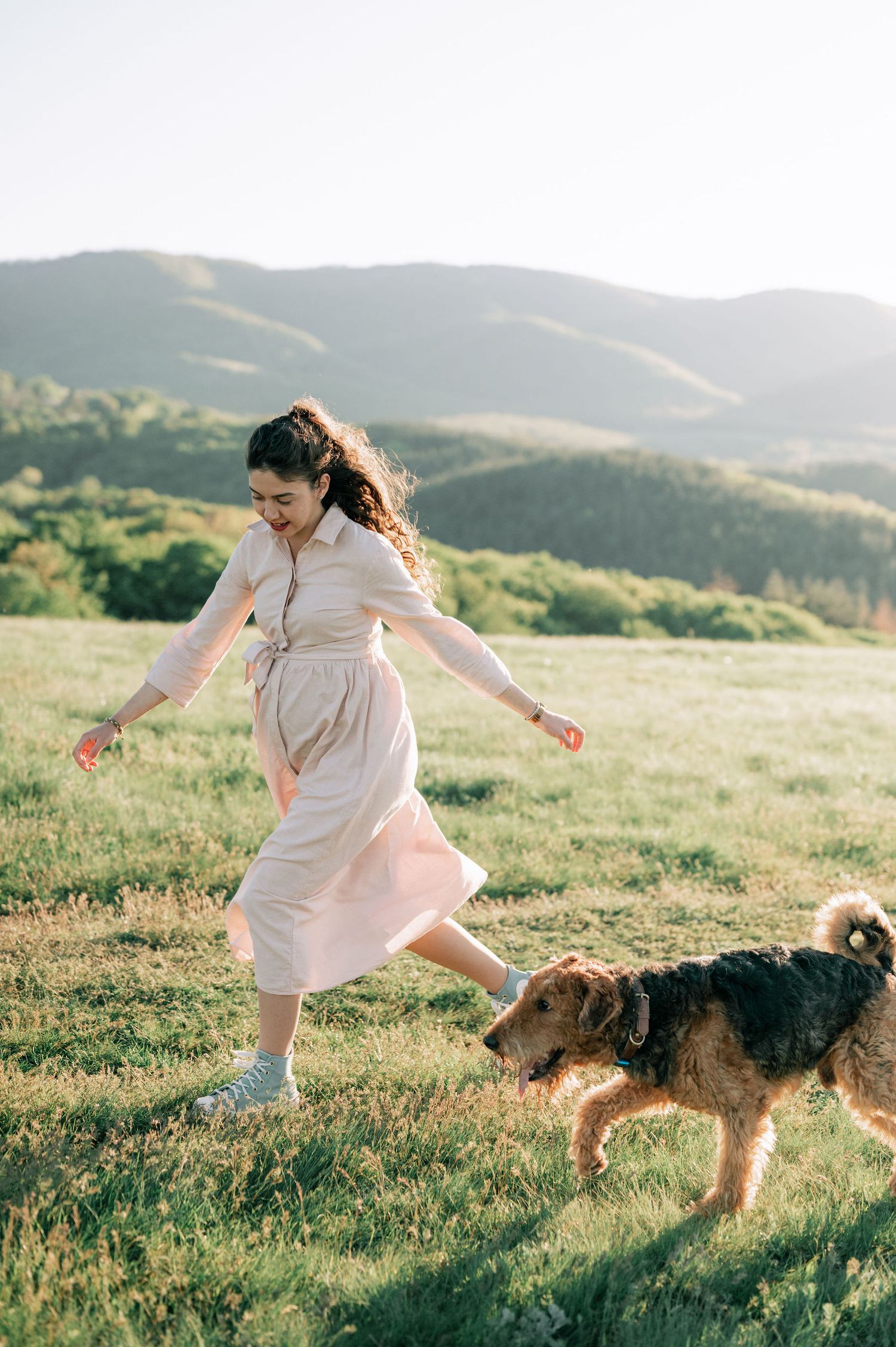 Person in flowy dress twirling joyfully with their Airedale Terrier dog in a sunlit mountain meadow.